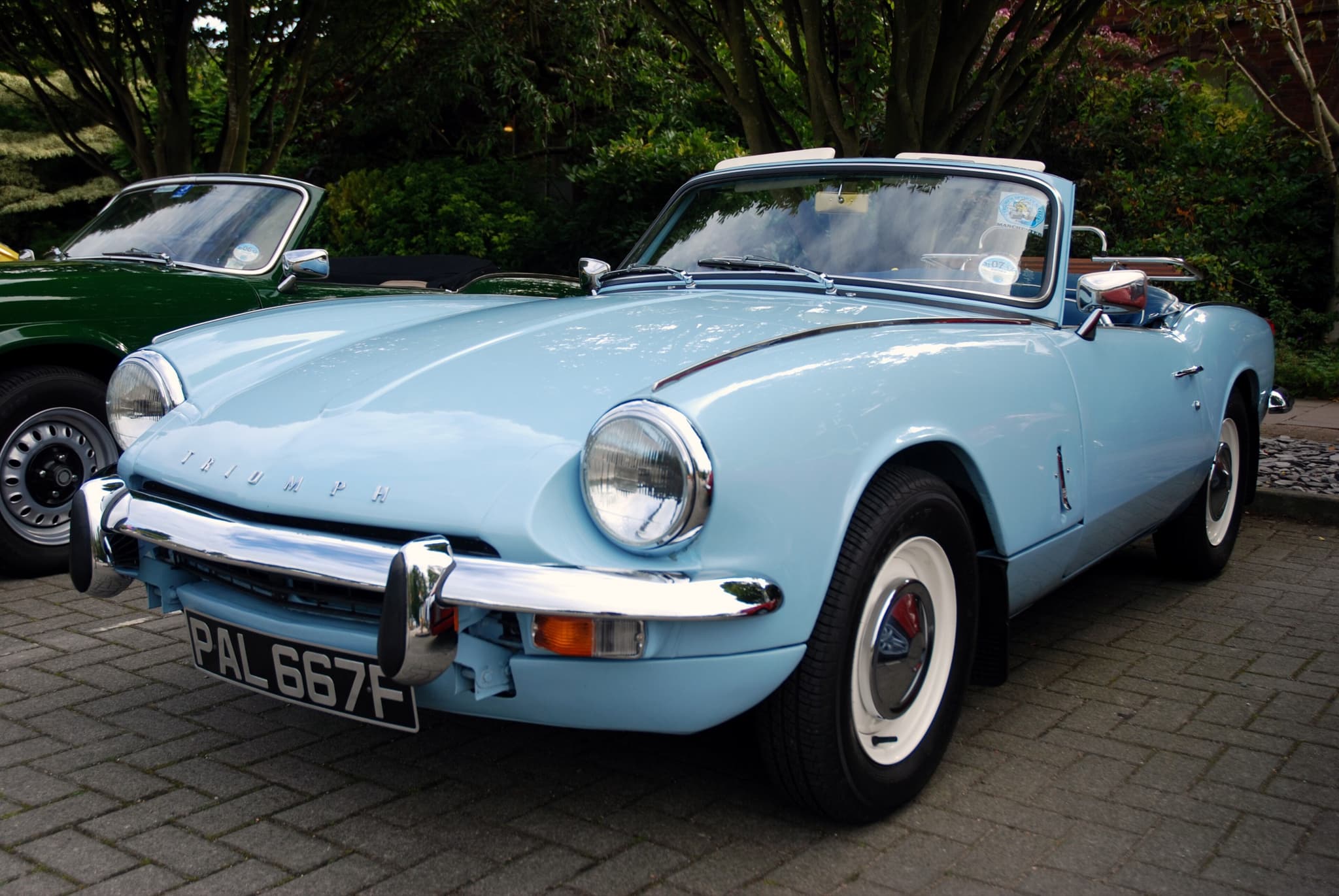 A pale blue Spitfire Mk III with white-painted wheels and its hood folded, parked on block paving. Opens a larger view