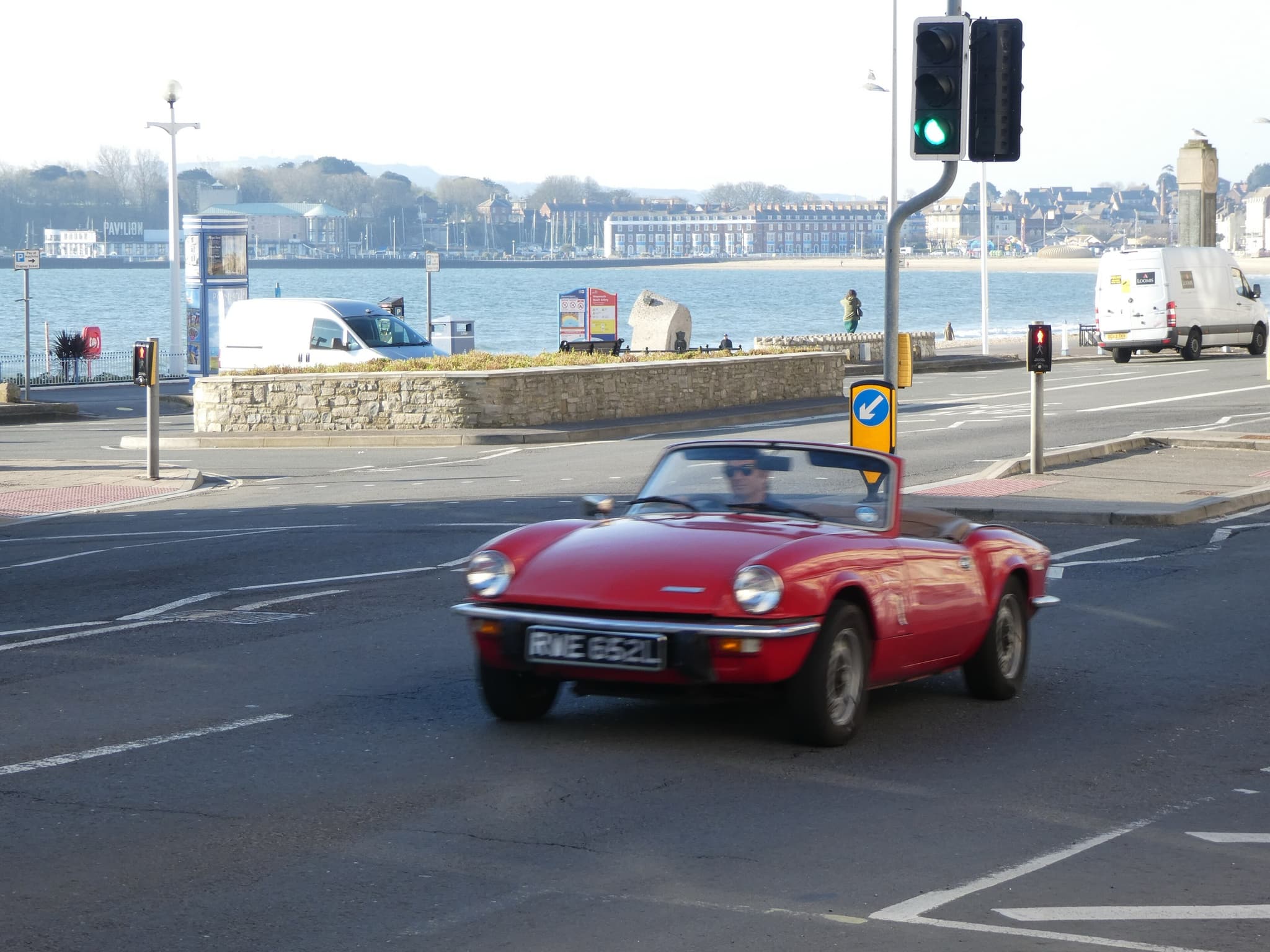 A red open Spitfire turning at a seafront road junction, with the bay and town behind.