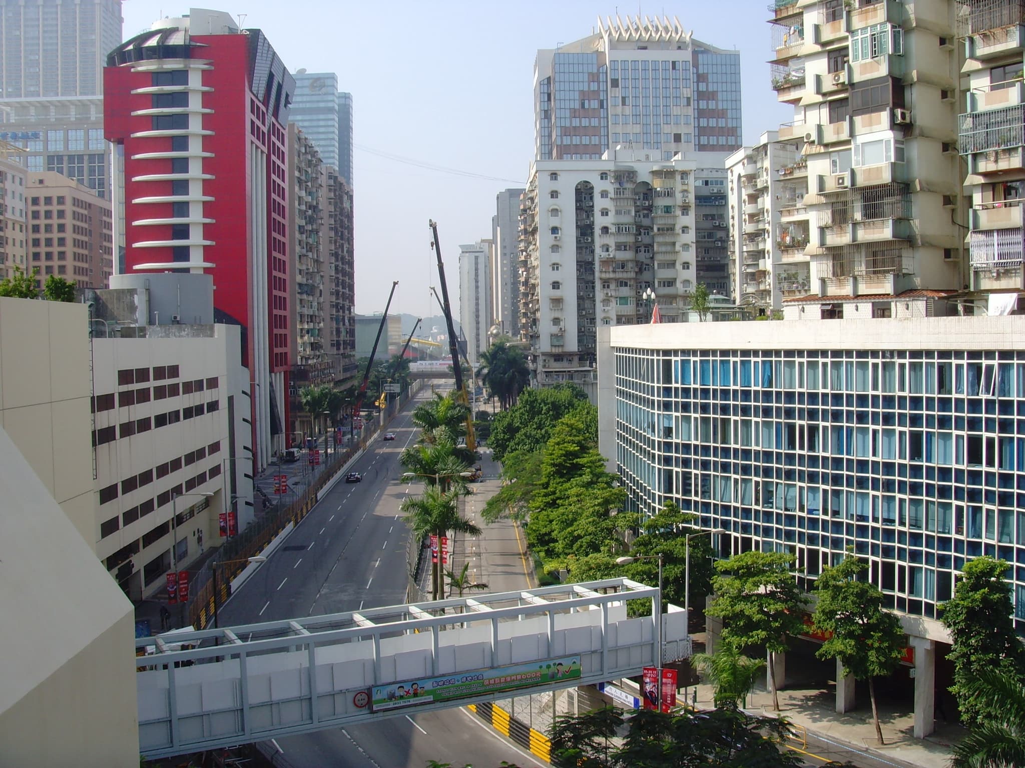 A city dual carriageway lined with barriers and striped kerbs, a footbridge crossing it below tall buildings.