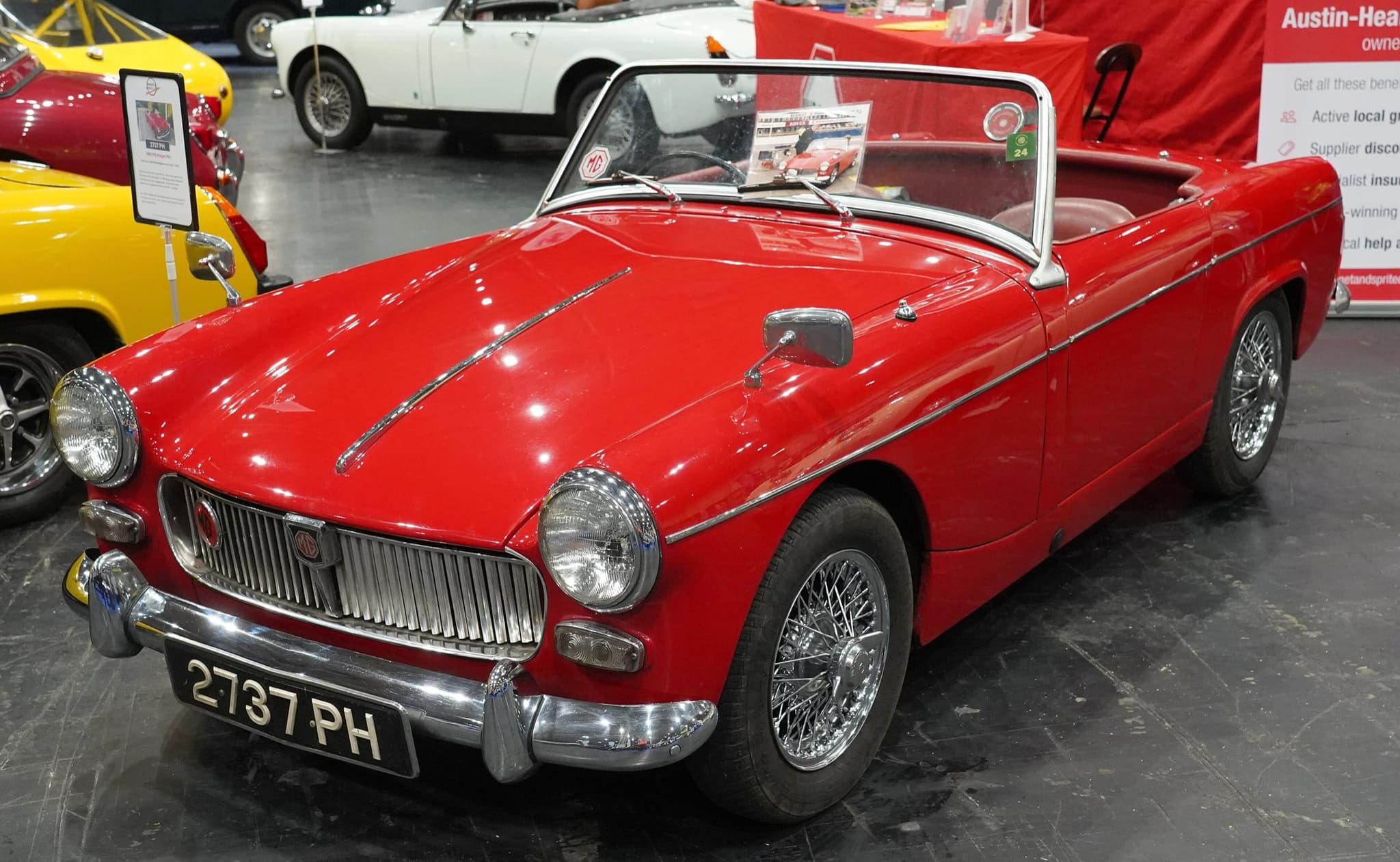 A red open two-seater with a vertical-slat grille, a chrome strip down the bonnet and wire wheels, on a show stand beside other small sports cars. Opens a larger view