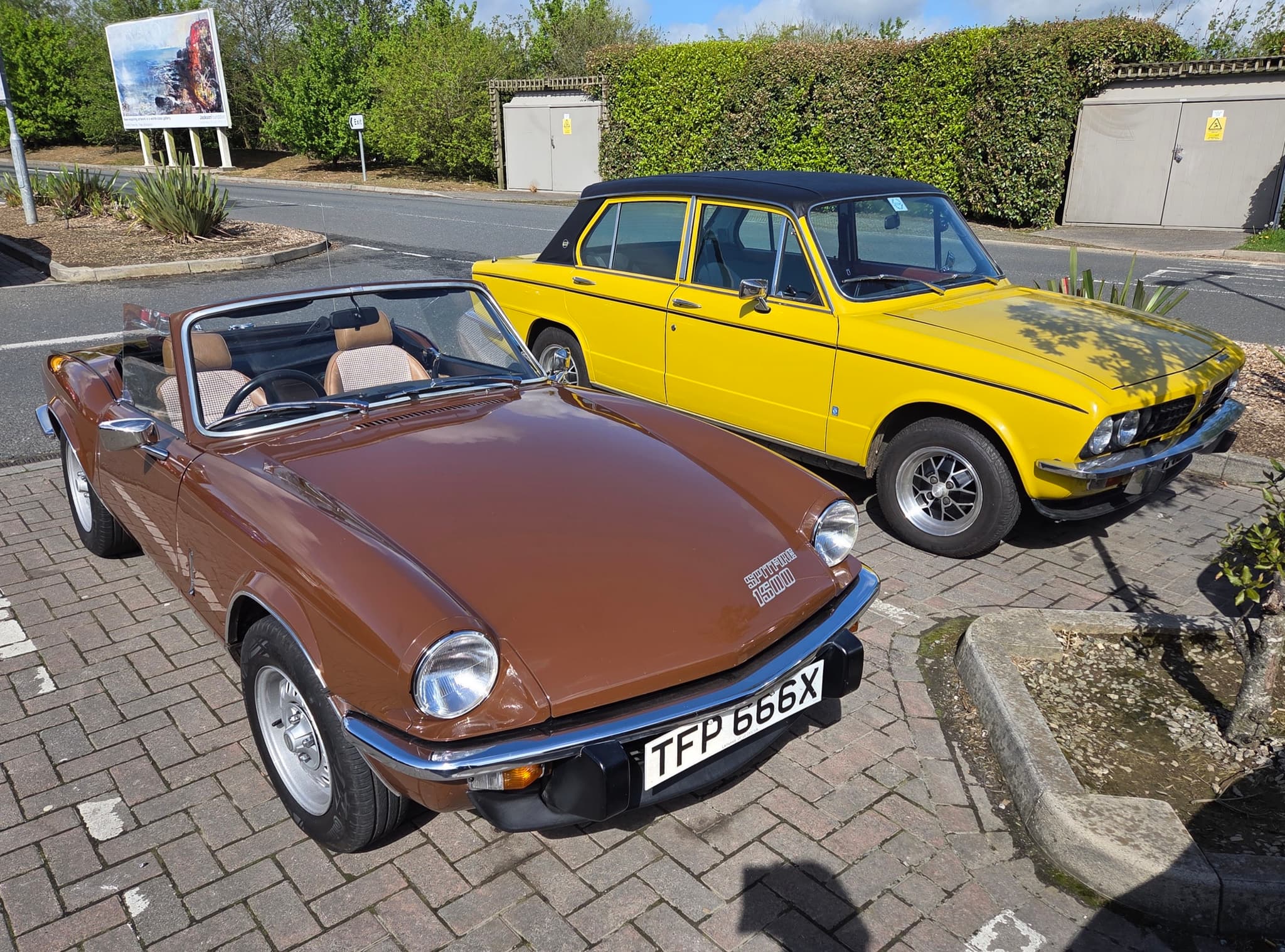 A brown Spitfire 1500 with its hood down beside a yellow Triumph saloon in a car park. Opens a larger view