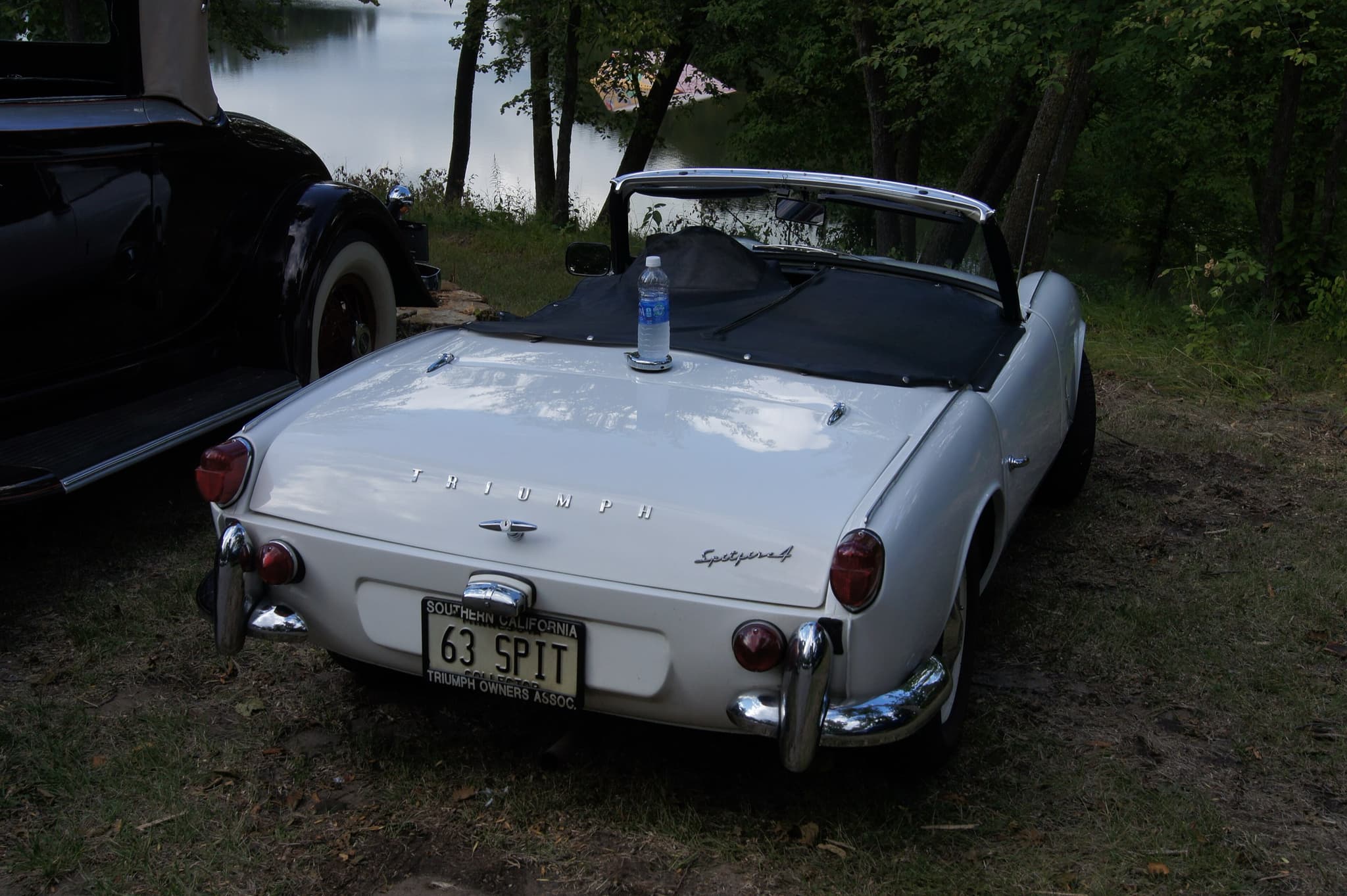 The tail of a white Spitfire 4 with chrome quarter-bumpers, parked on grass under trees beside a lake. Opens a larger view