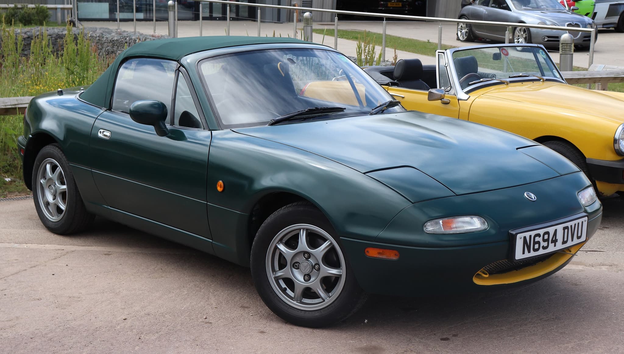 A dark green two-seater with its green hood up and pop-up headlamps closed, parked beside a yellow roadster.