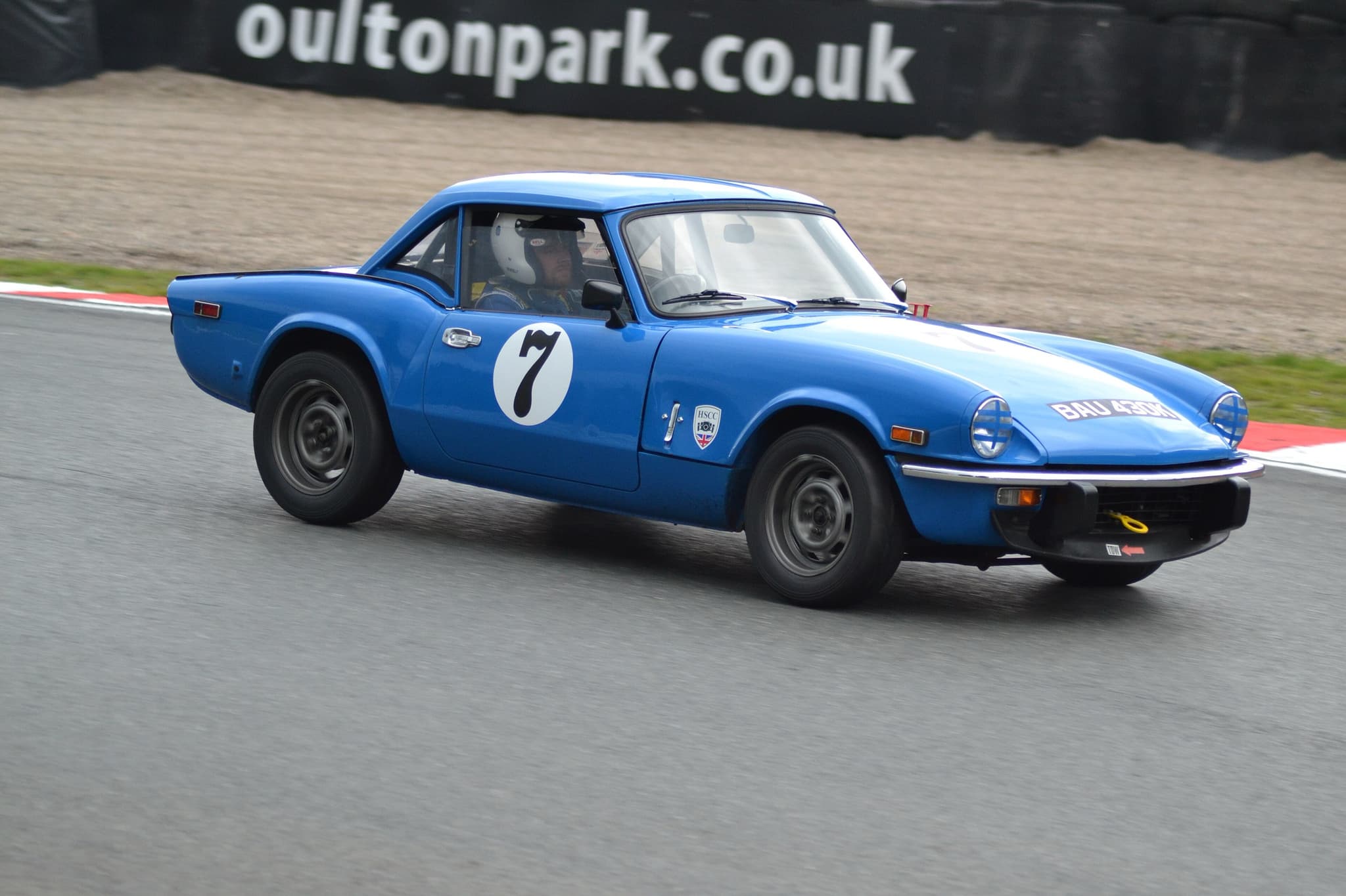 A blue Spitfire with a hardtop and race number 7 cornering on a circuit beside a gravel trap. Opens a larger view