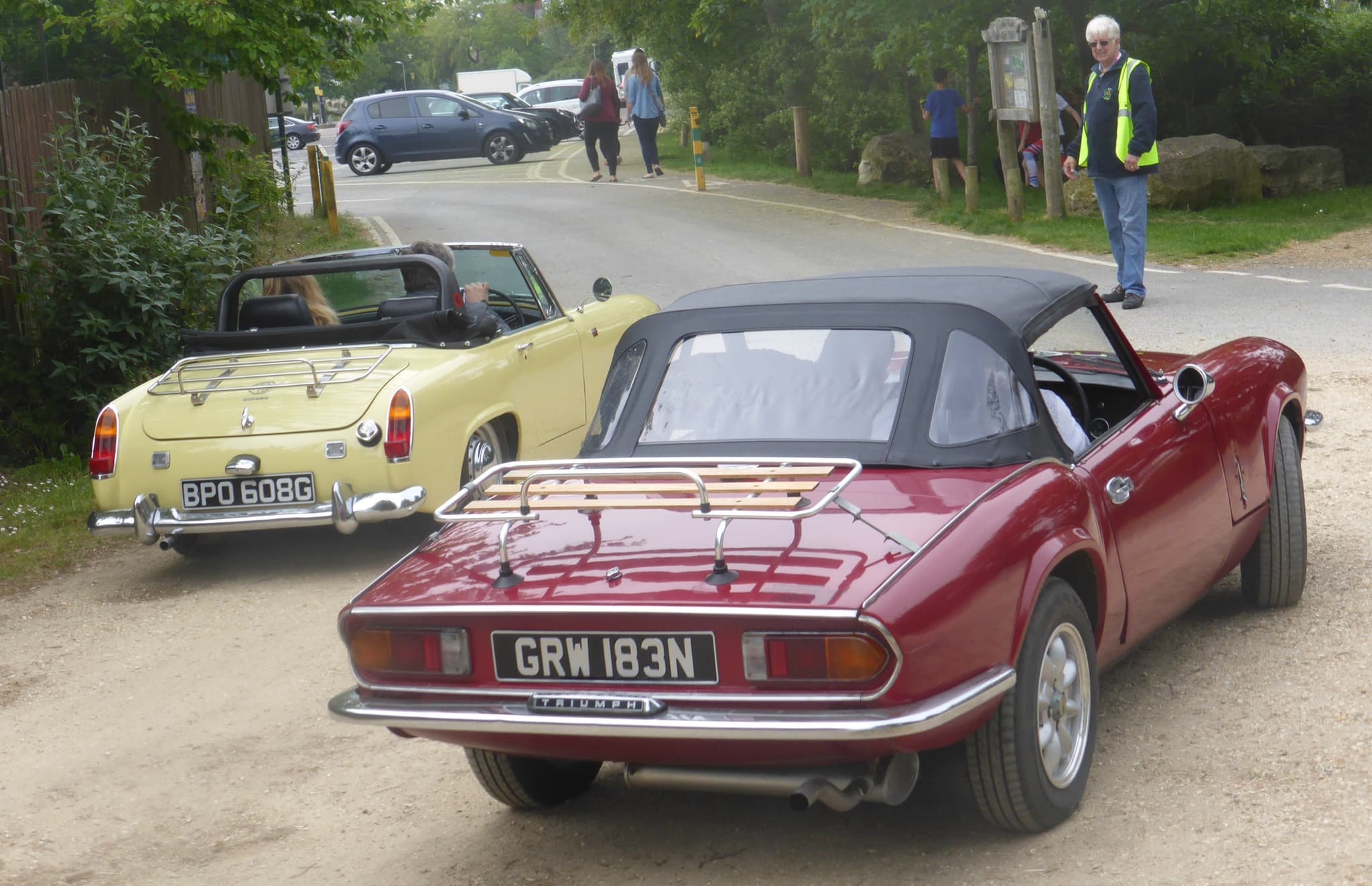 Two small open sports cars from behind on a gravel lane: a pale yellow roadster with its hood down and a dark red one with a black hood and a wooden luggage rack, a man in a high-visibility vest watching at the right. Opens a larger view