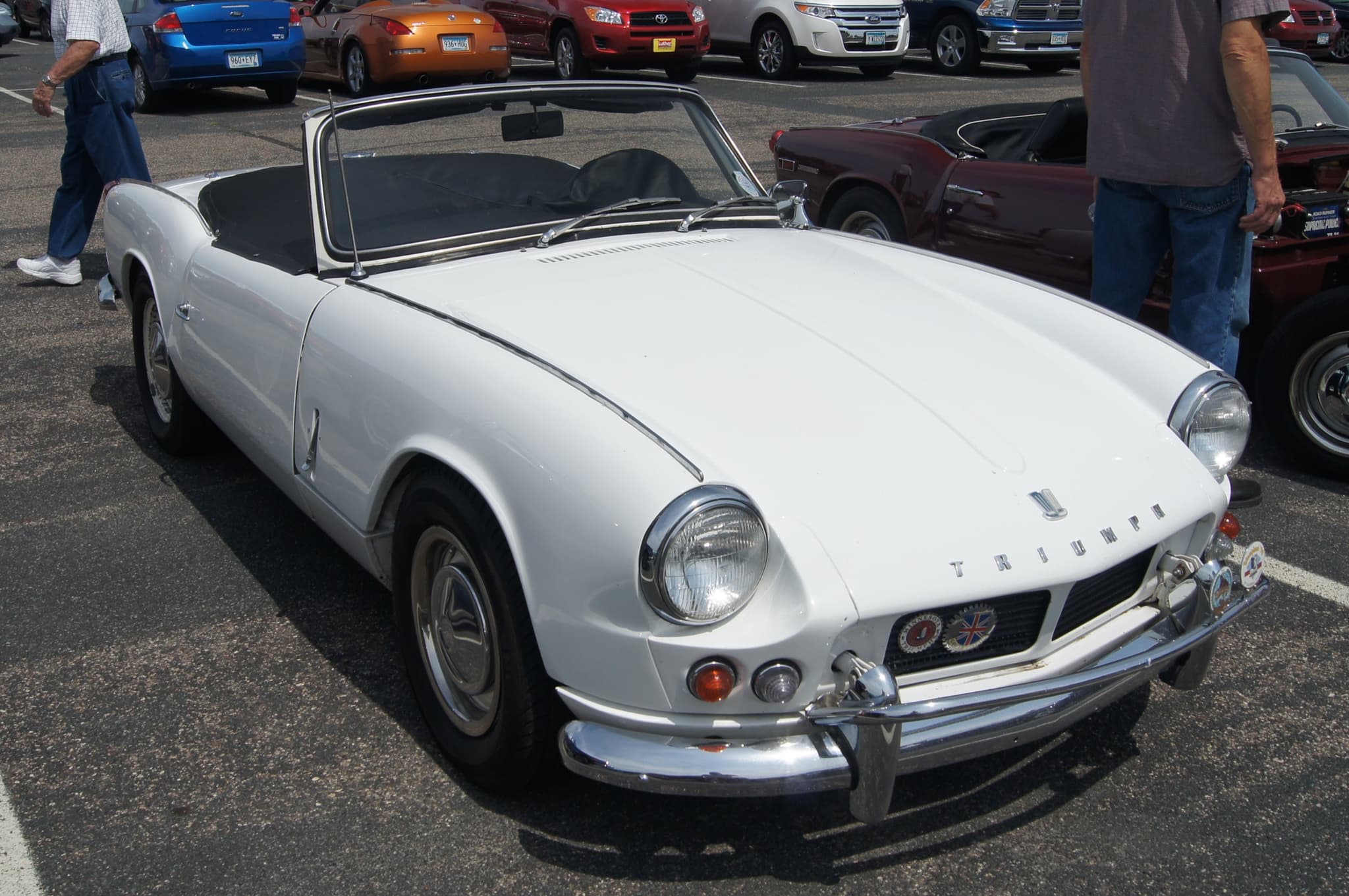 A white early Spitfire with its hood folded, chrome bumper and overriders, in a sunny car park among modern cars.