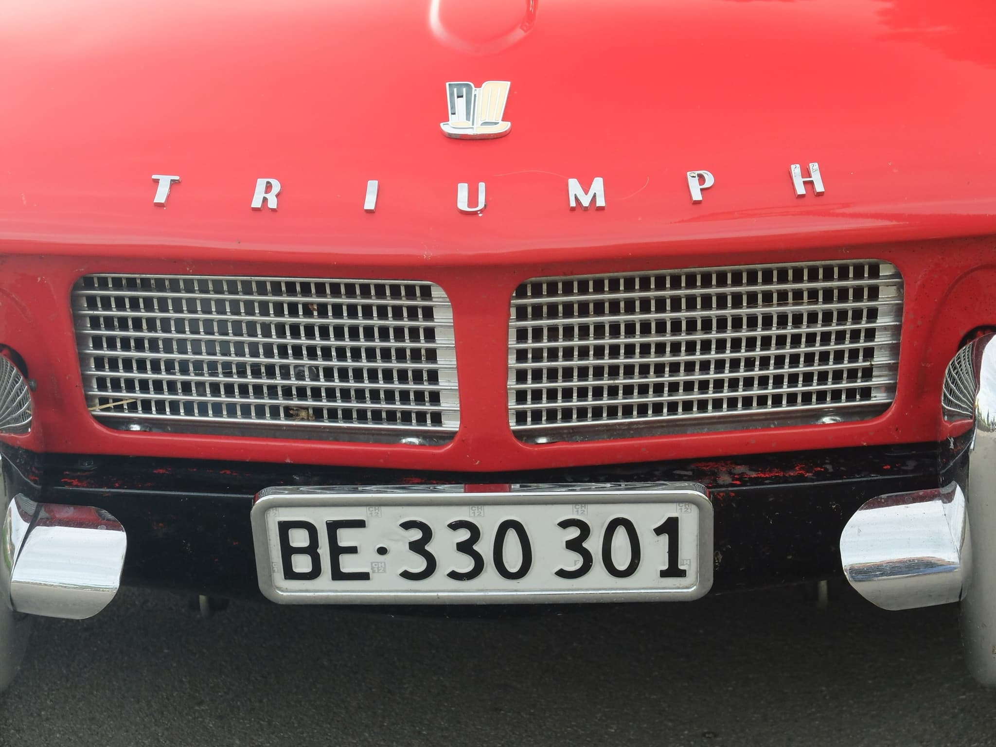 Close-up of a red Spitfire 4 nose: bonnet badge, chrome TRIUMPH letters and two fine-mesh grille halves. Opens a larger view