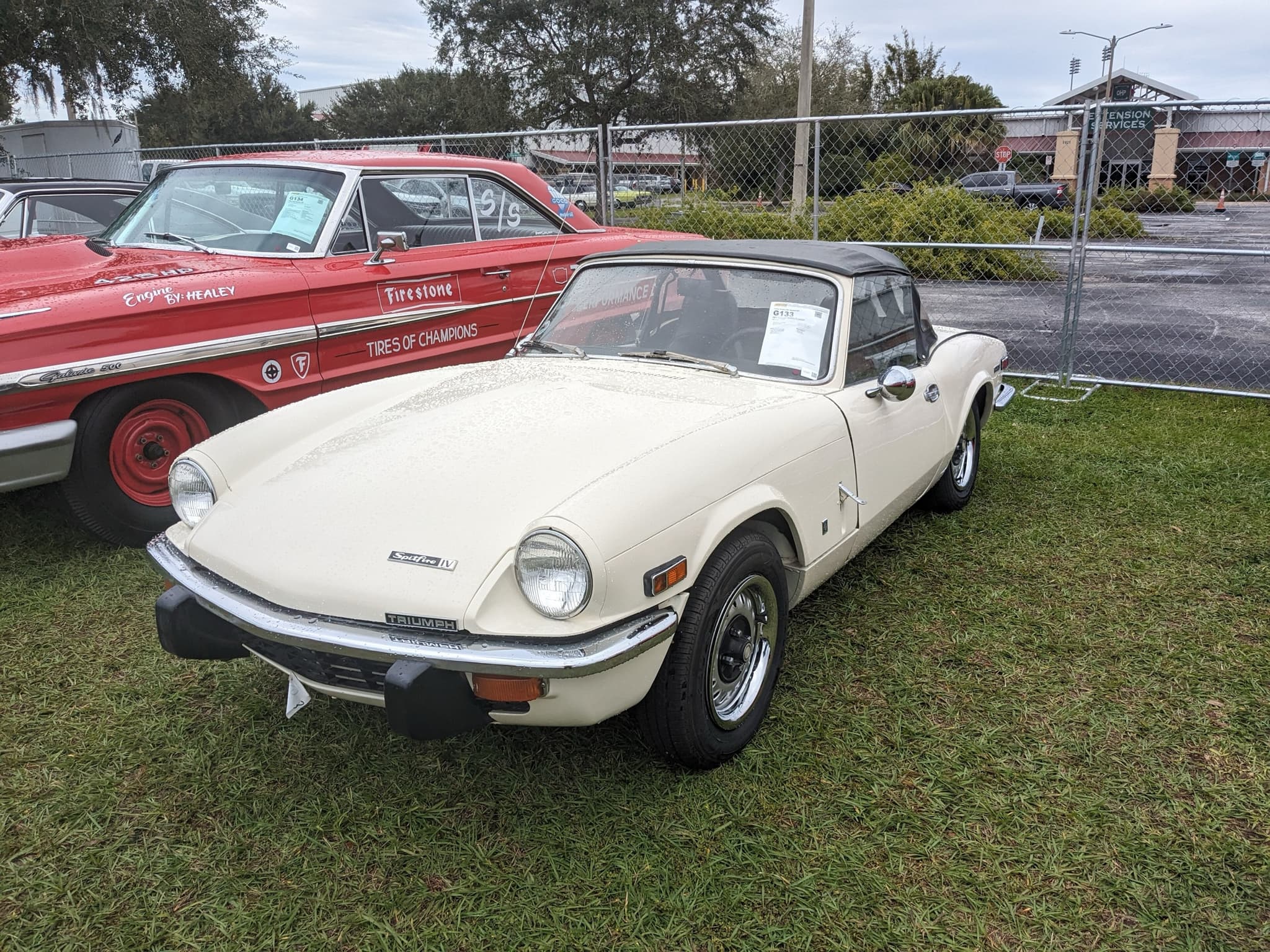 A cream Spitfire with its black hood up, 'Spitfire IV' bonnet badge, amber side lamps and black rubber overriders, on wet grass.