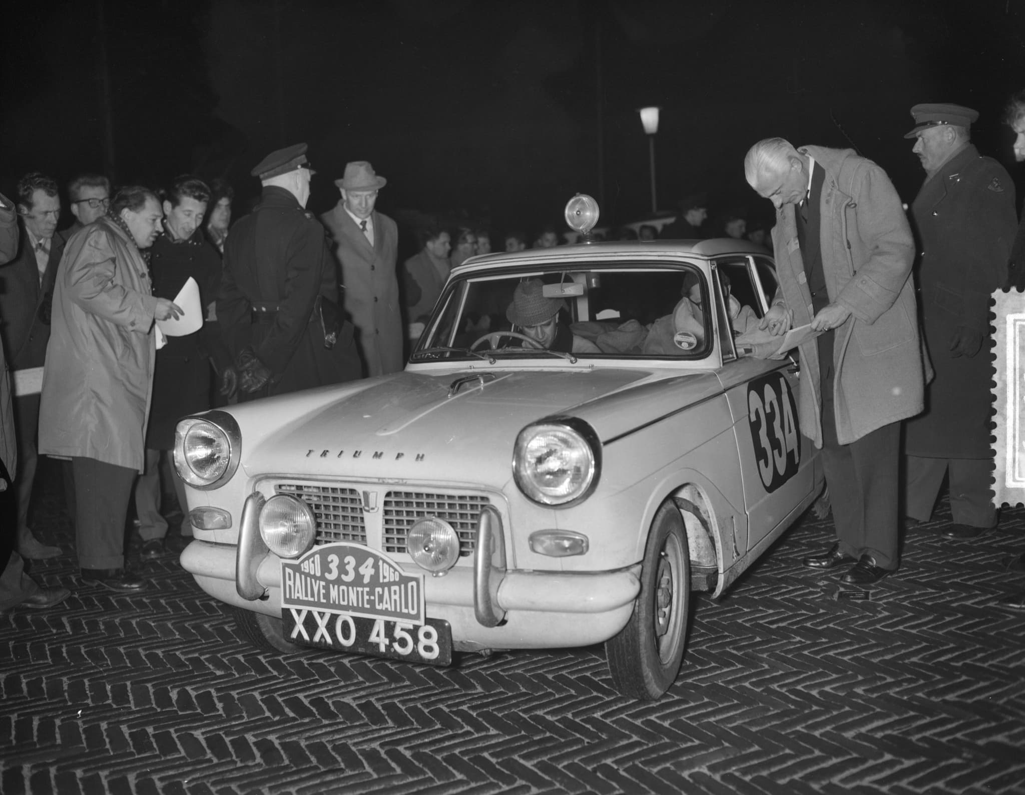 A rally-plated Triumph Herald saloon, number 334, with spot lamps, among officials and onlookers on a brick street at night.