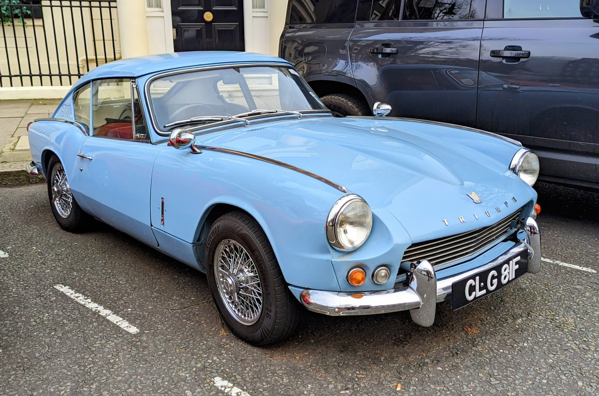 A pale blue fastback coupé with wire wheels and a low chrome bumper, parked at a kerb beside a dark modern car.