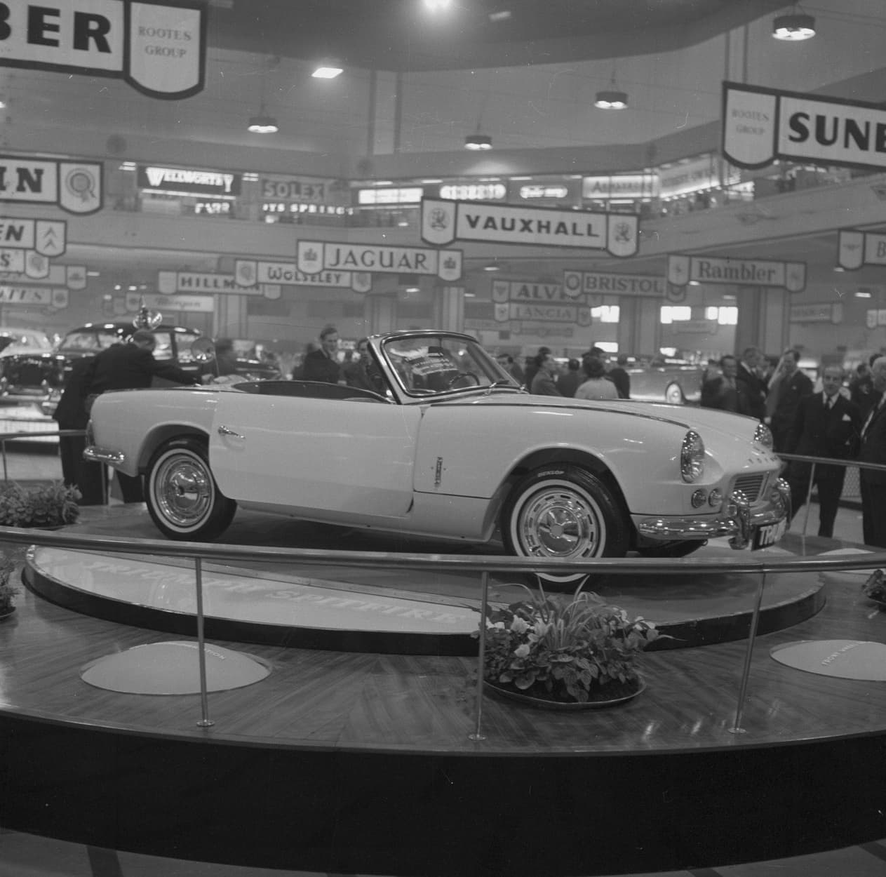 A black-and-white photograph of a Spitfire 4 from the front three-quarter on a lit turntable at a motor show, a man leaning into its cockpit and stand signs overhead.