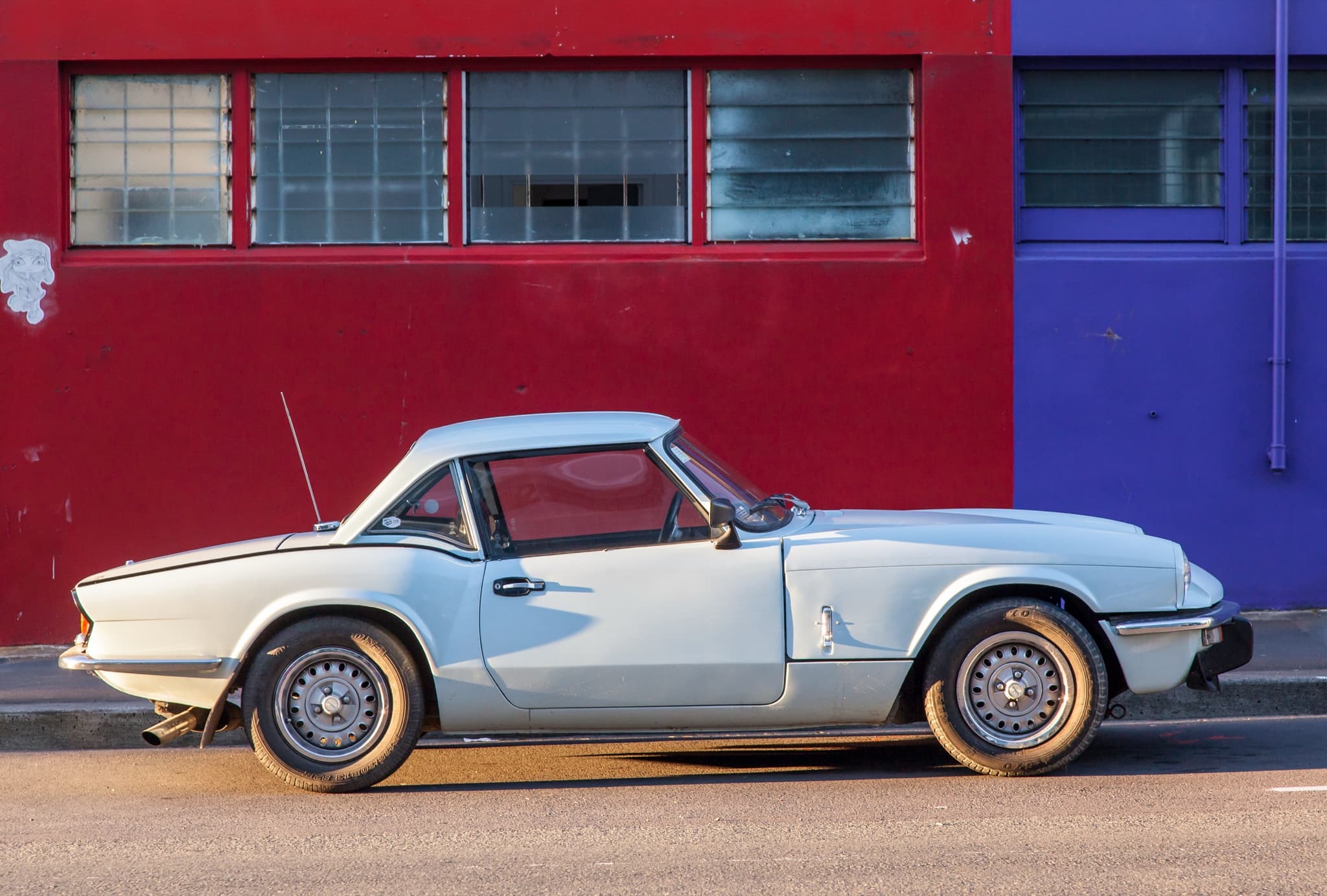 A pale blue Spitfire 1500 with a hardtop in exact profile against red and purple painted walls.