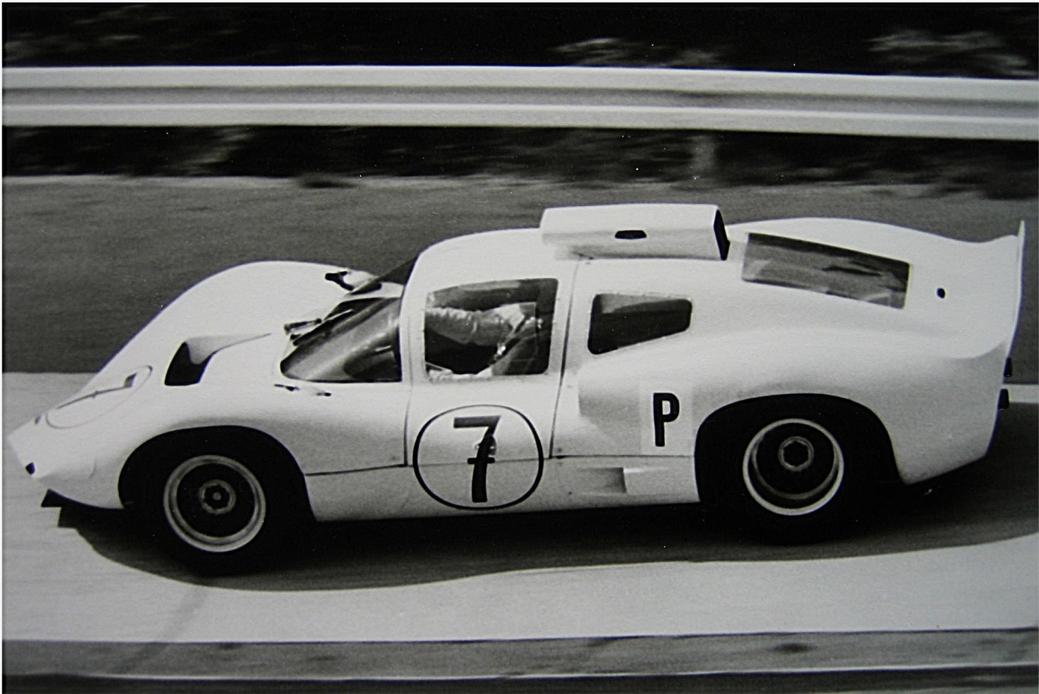 A white closed racing car with a tall rear wing and the number 7, cornering past a steel barrier, the driver visible.