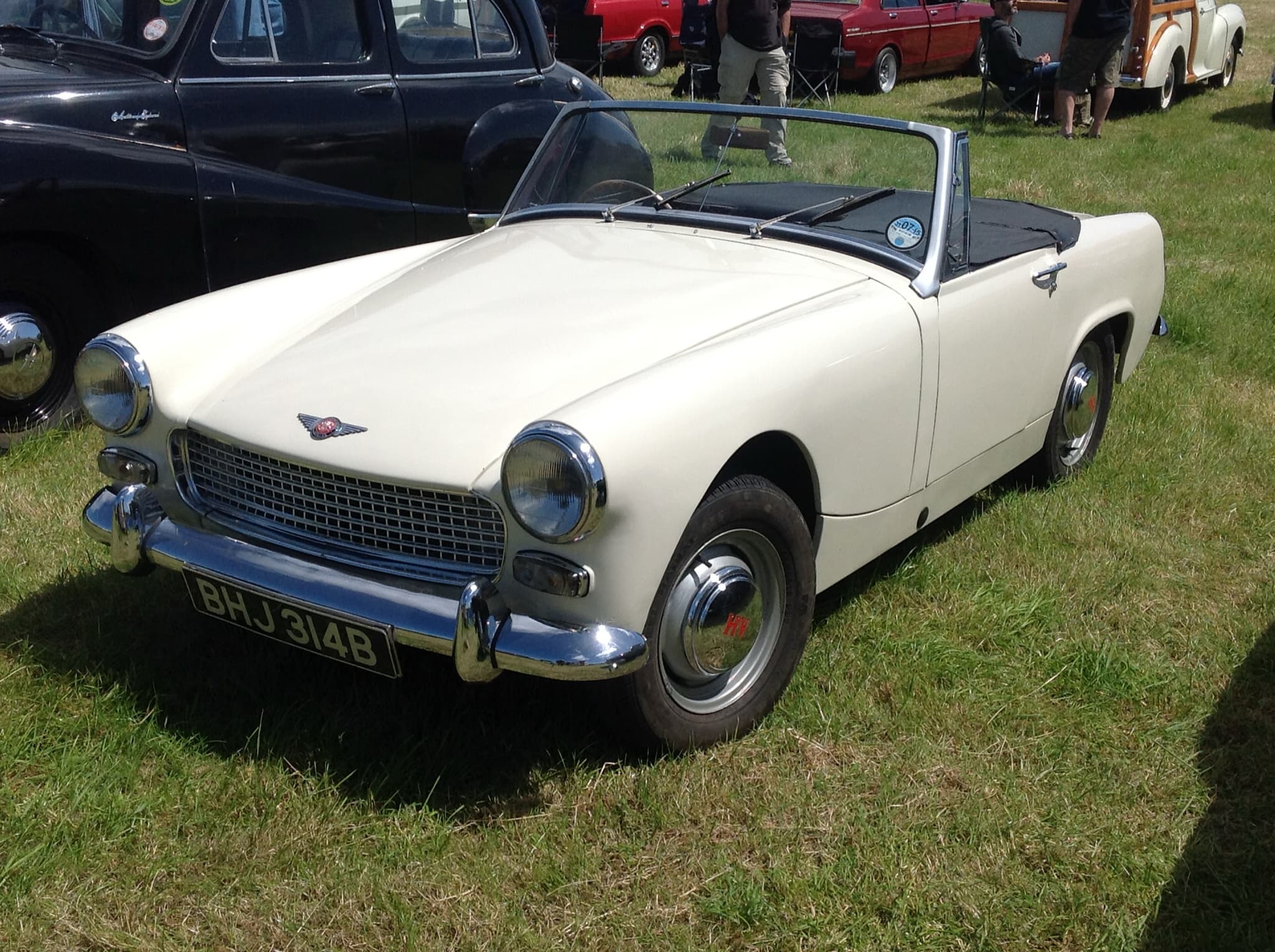 A cream open two-seater with round headlamps and a chrome bumper, parked on grass in front of a black saloon and other old cars. Opens a larger view
