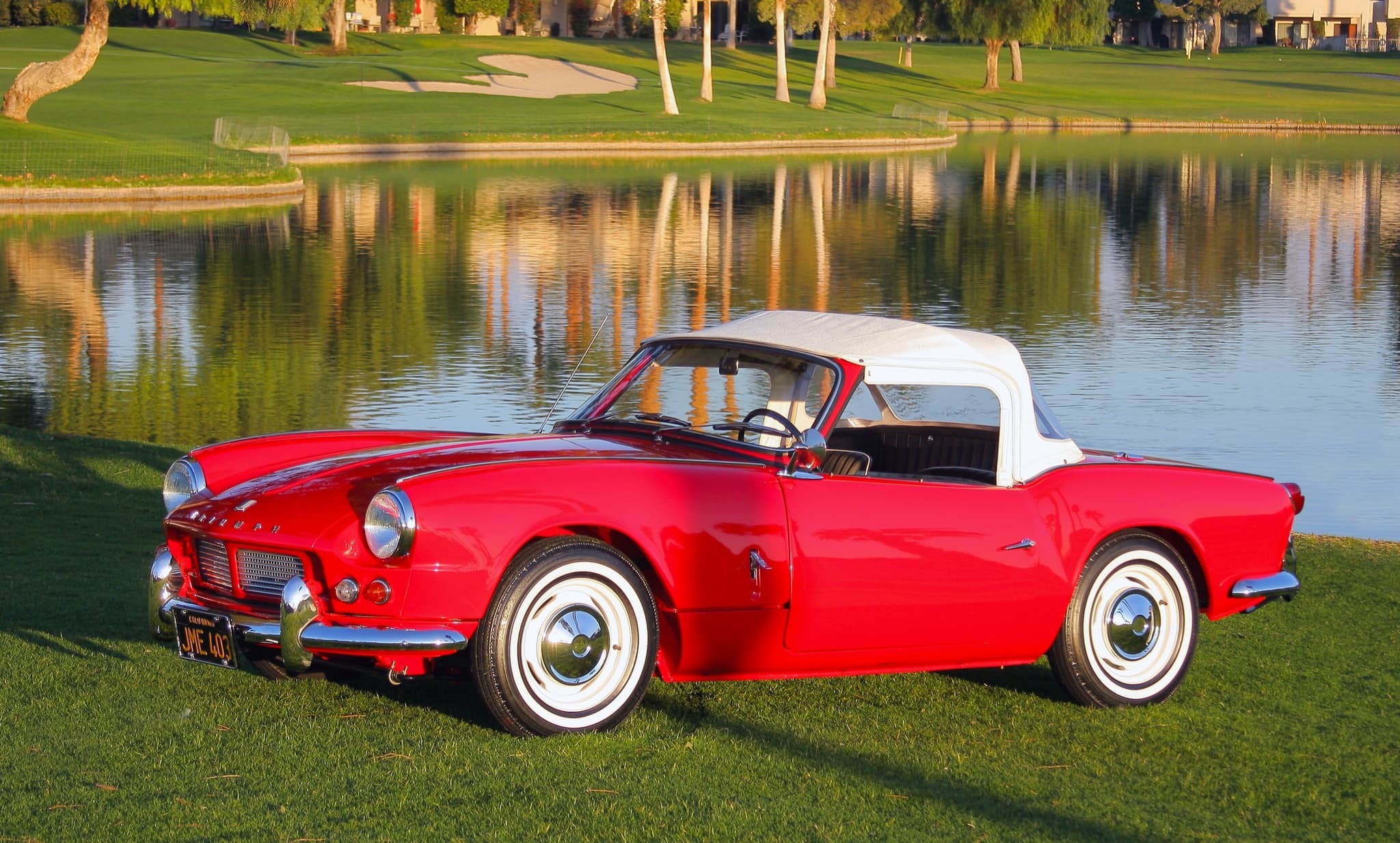 A red Spitfire 4 with a white hood and whitewall tyres on grass beside a still lake. Opens a larger view