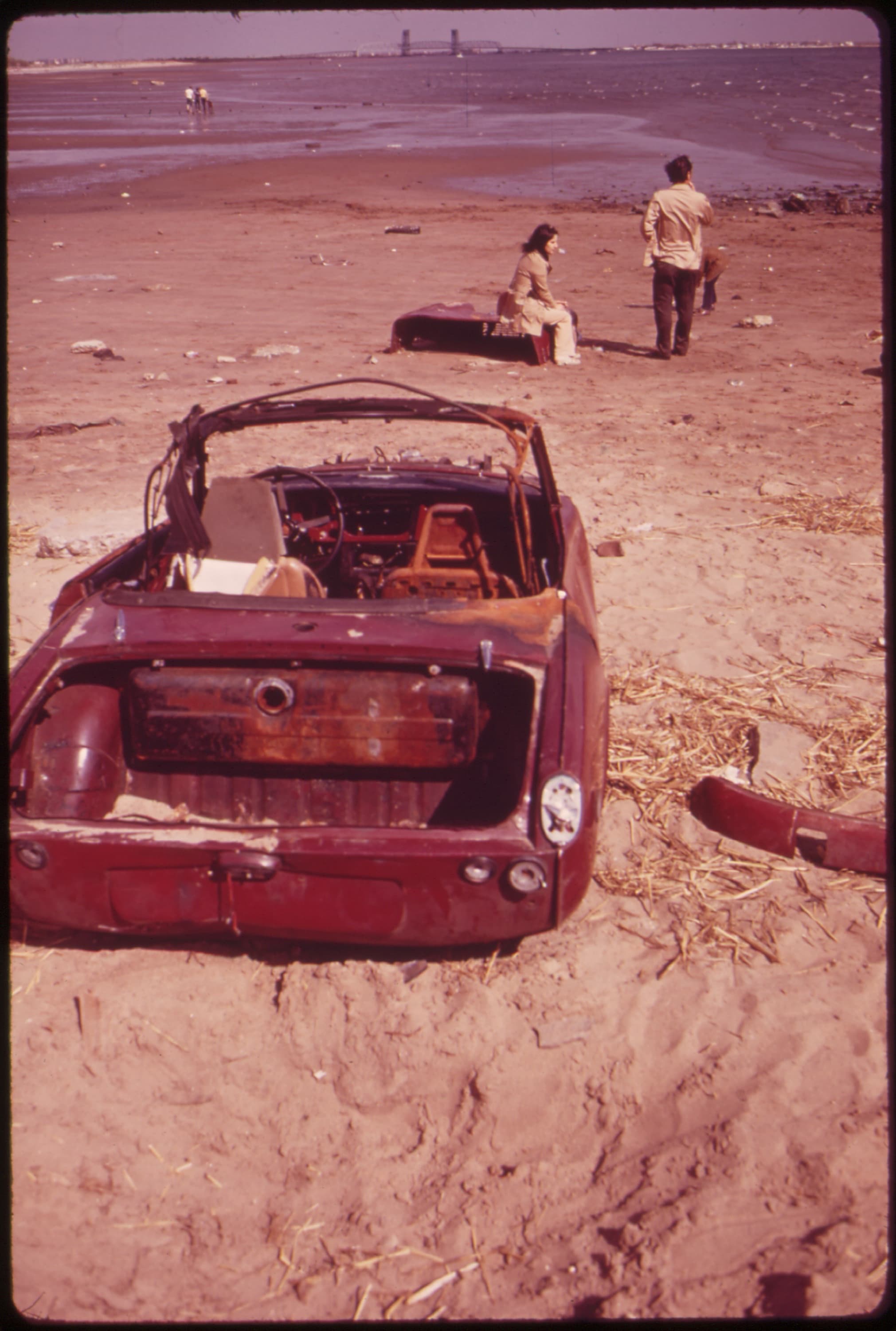 A stripped, rusting round-tail Spitfire sunk in beach sand, its rear bumper lying beside it, in faded period colour. Opens a larger view