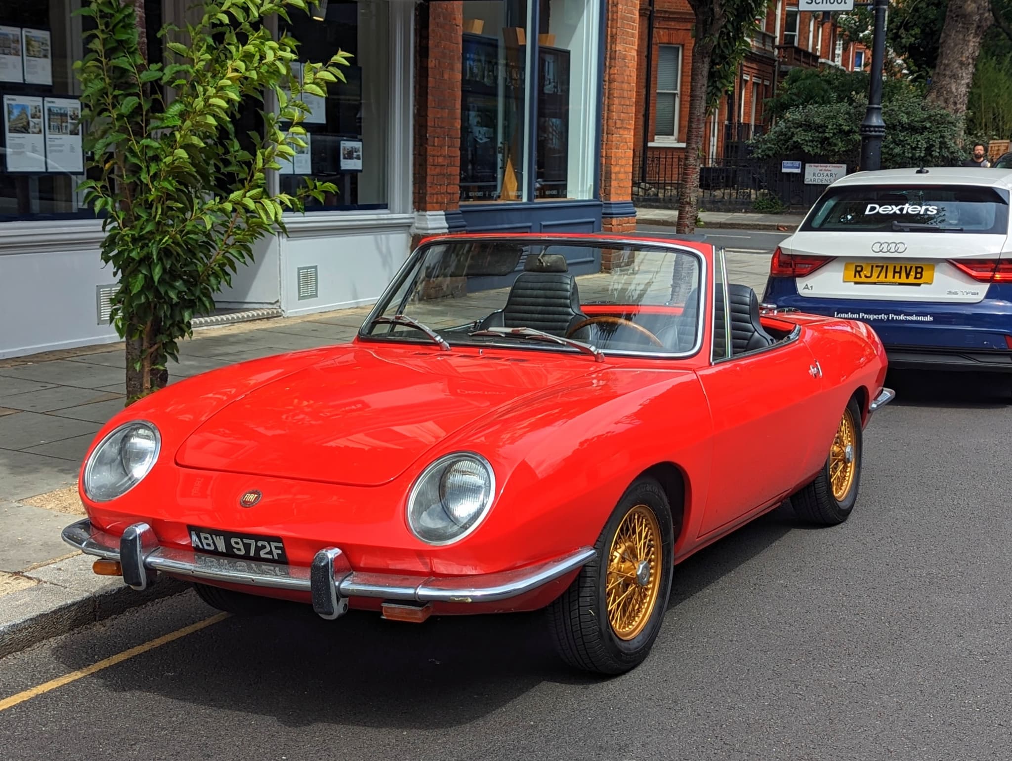 A small red Italian roadster with faired-in headlamps, slim chrome bumpers and gold wire wheels, parked at a city kerb. Opens a larger view