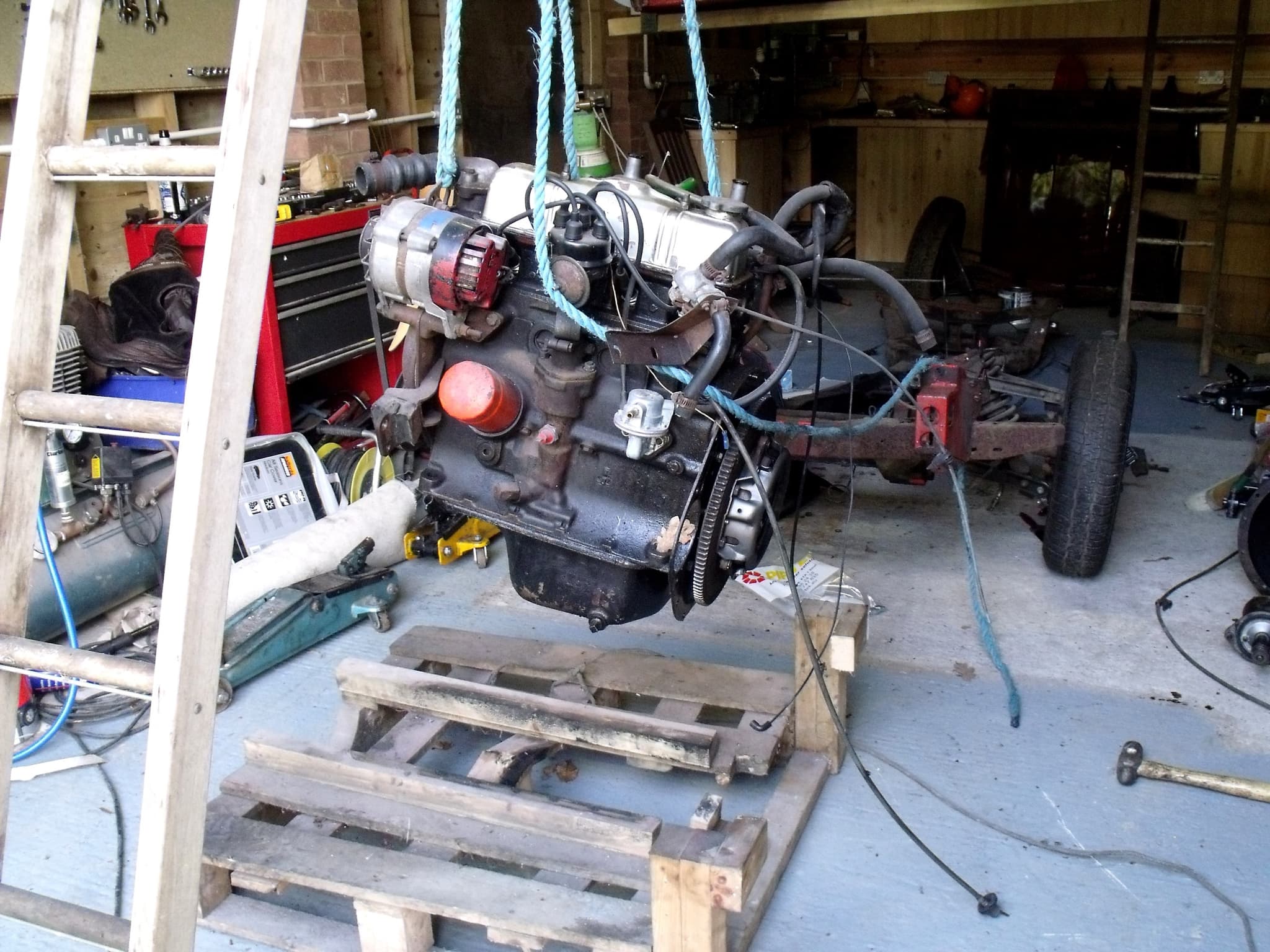 A four-cylinder engine hanging on blue ropes over a wooden pallet, with a bare chassis and a wheel behind it in a cluttered garage.