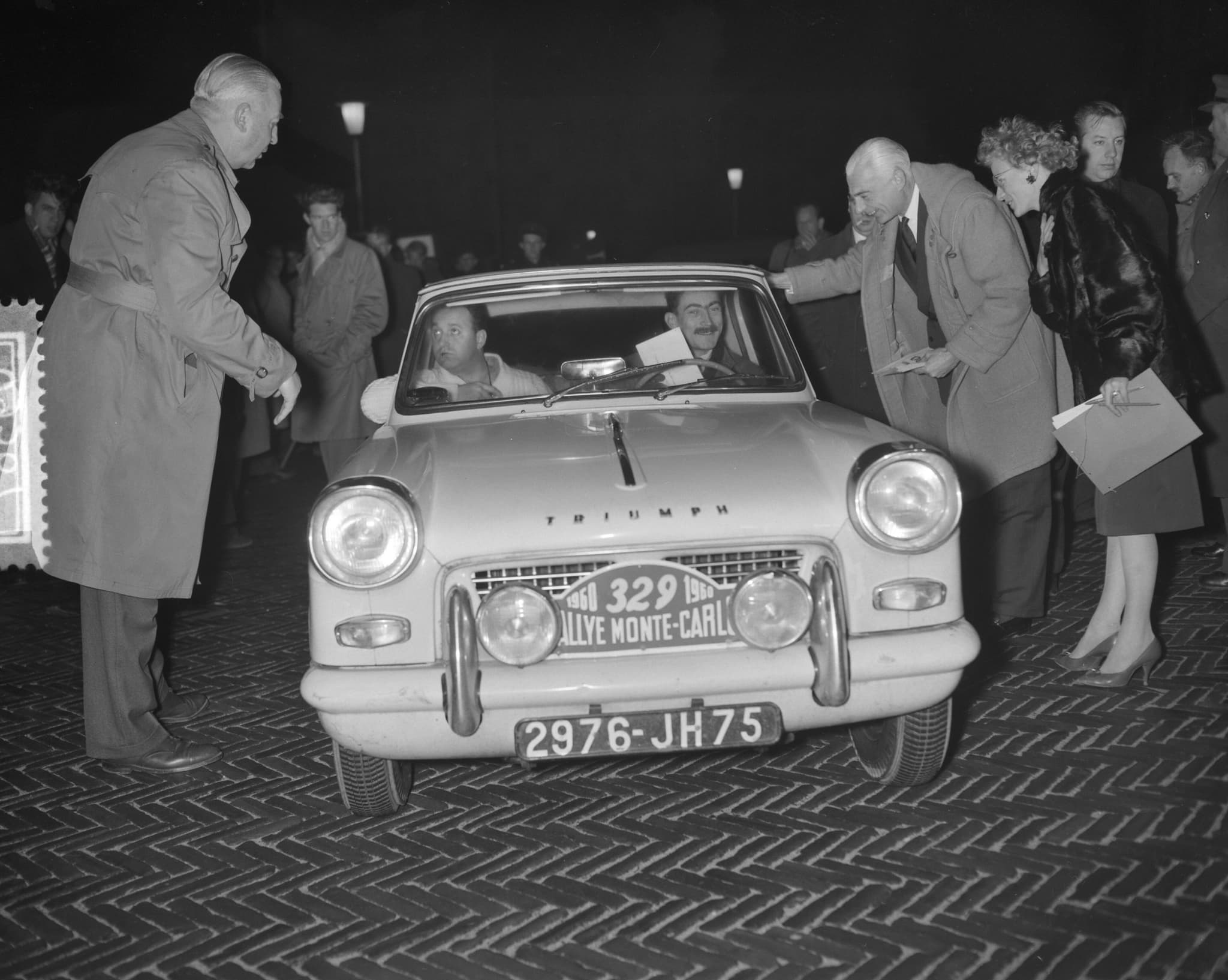 A rally-plated Triumph Herald saloon, number 329, its crew aboard, as officials lean in at the window at night.