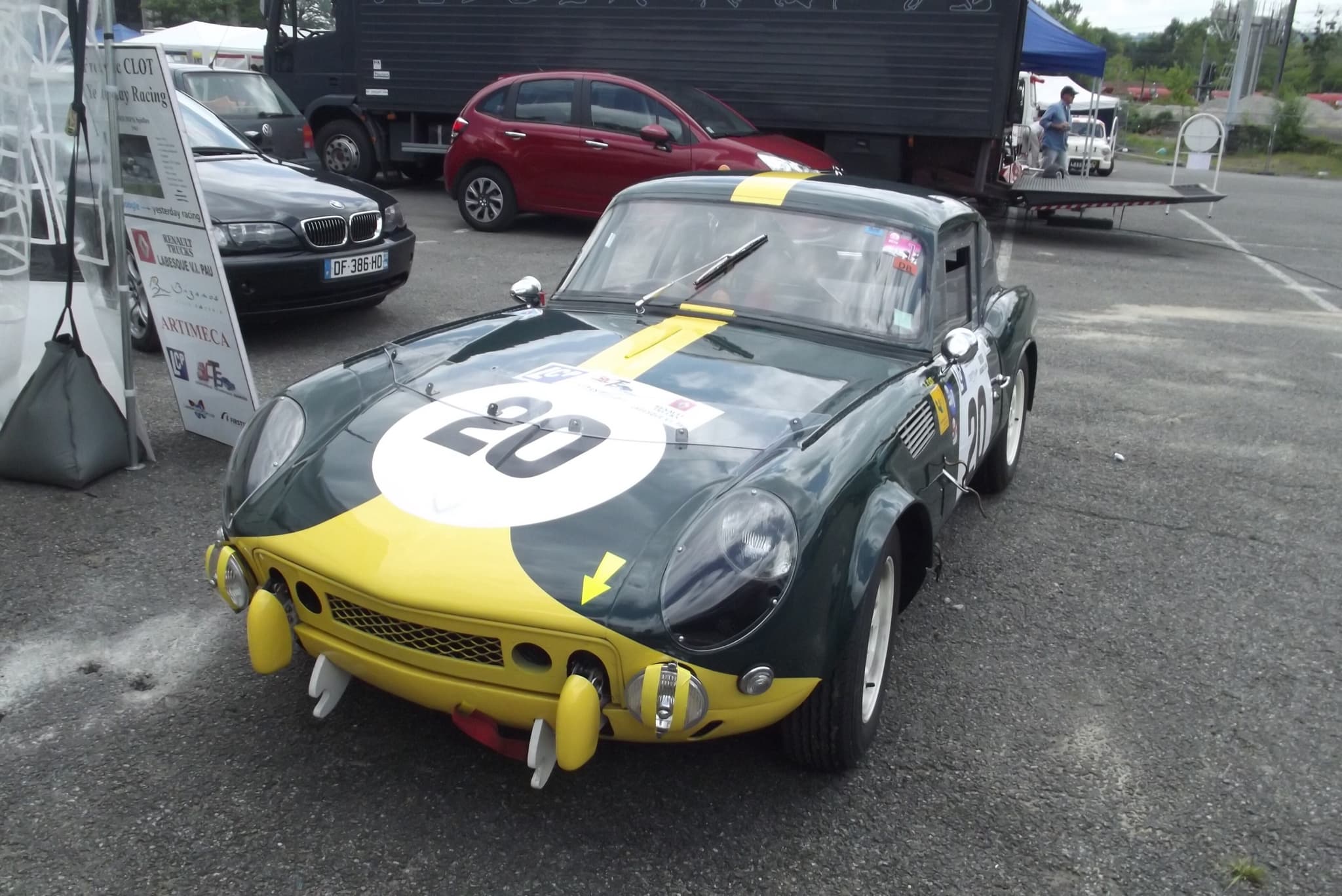 A dark green fastback Spitfire race car with a yellow nose and race number 20 in a paddock. Opens a larger view