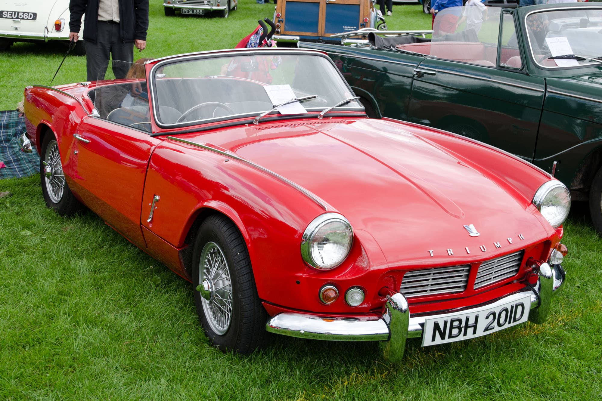A red Spitfire 4 Mk II with the hood down and wire wheels, parked on a show field among classics. Opens a larger view