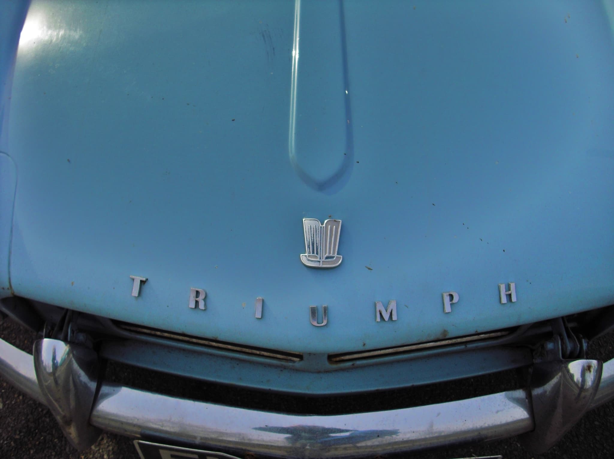 Close-up of a pale blue bonnet with its central ridge, badge and chrome TRIUMPH letters above the bumper. Opens a larger view