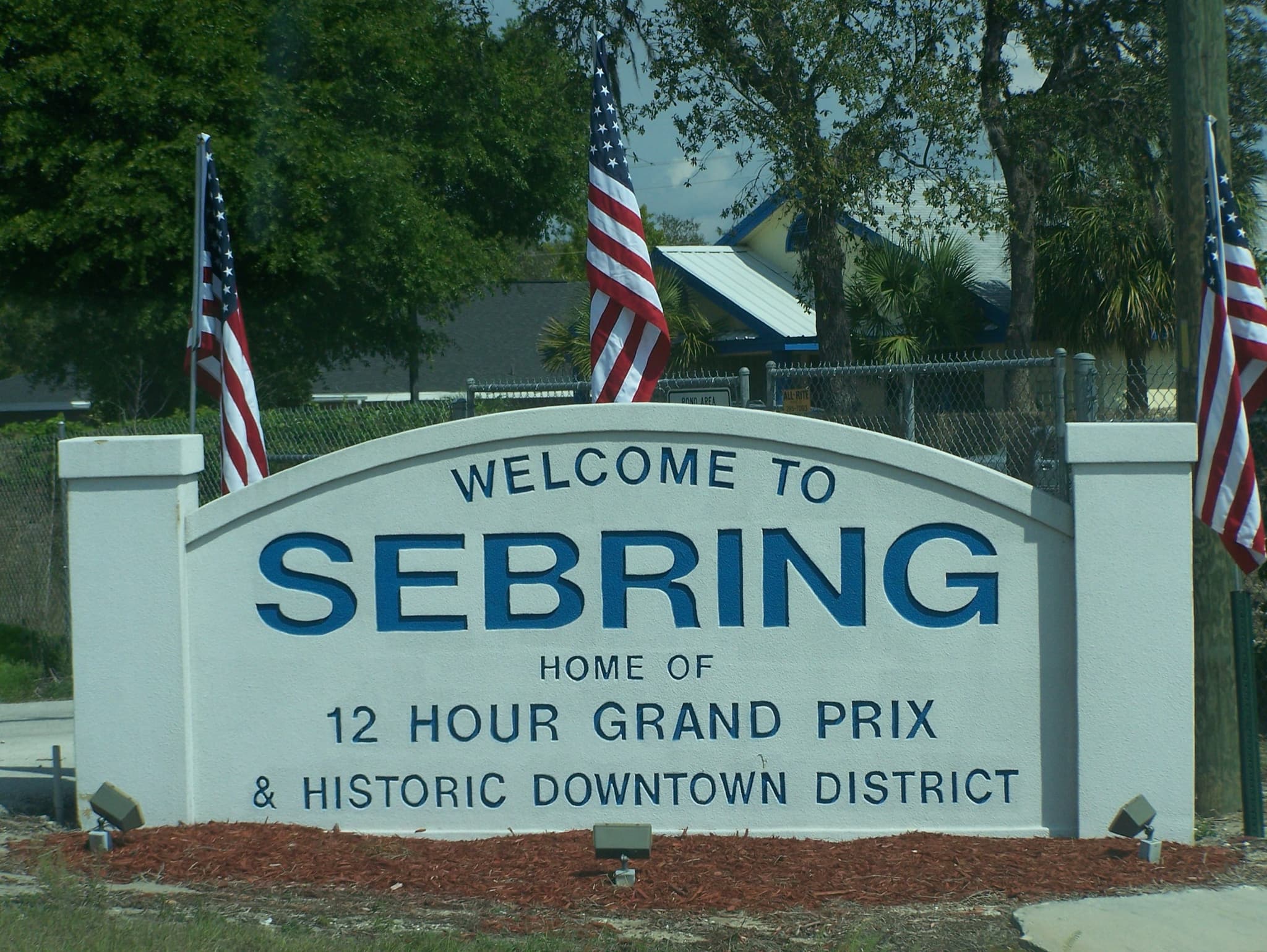 A white town sign reading Welcome to Sebring, home of 12 Hour Grand Prix, with three American flags.