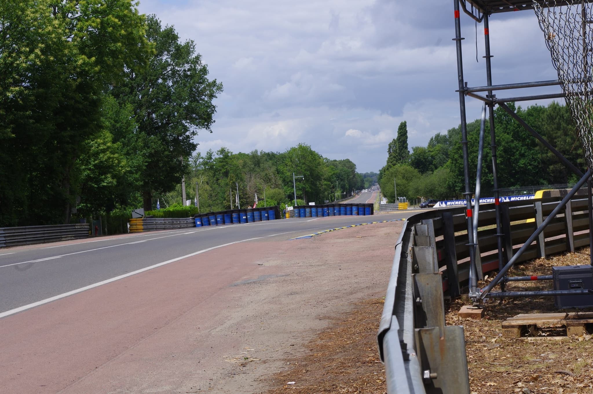An empty straight road between trees, with steel barriers, a red run-off strip and a chicane.