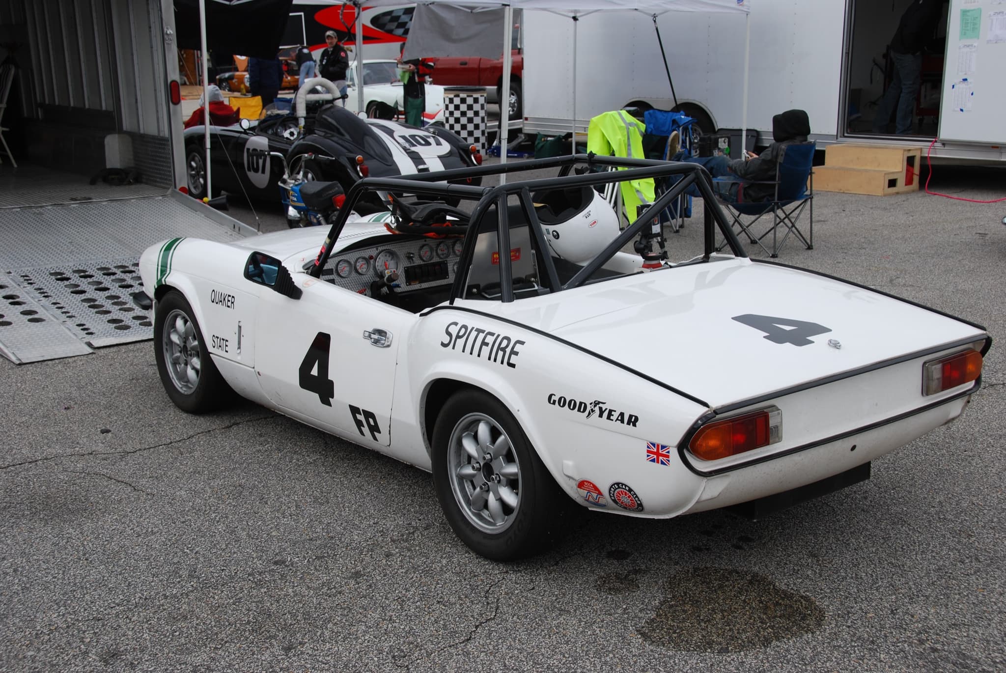 A white Spitfire race car numbered 4, with a roll cage, parked among trailers and tents in a race paddock. Opens a larger view