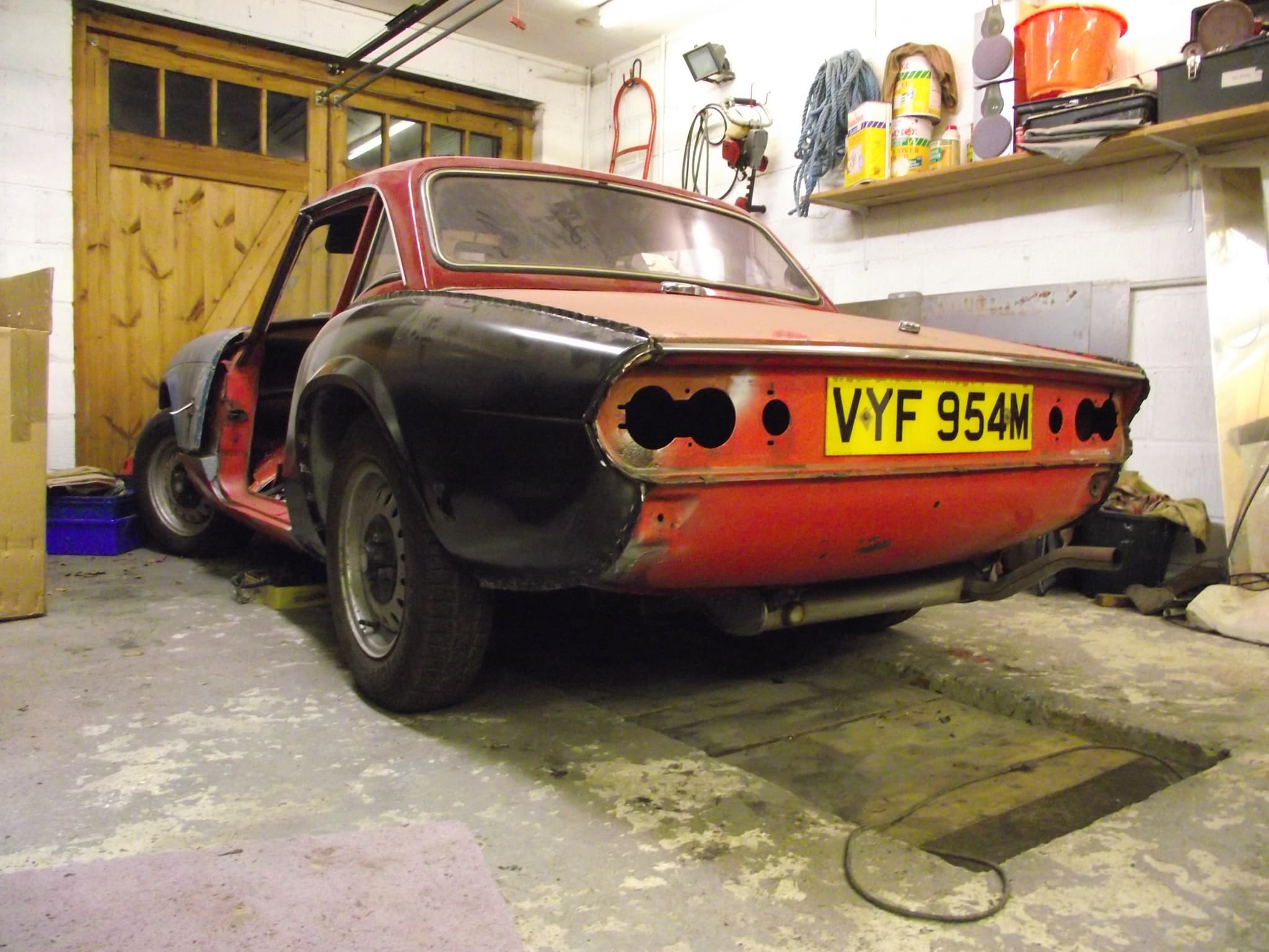 A red Spitfire with a hardtop seen low from behind in a garage, its rear lamps removed and its rear quarter black, shelves of tins beside it.