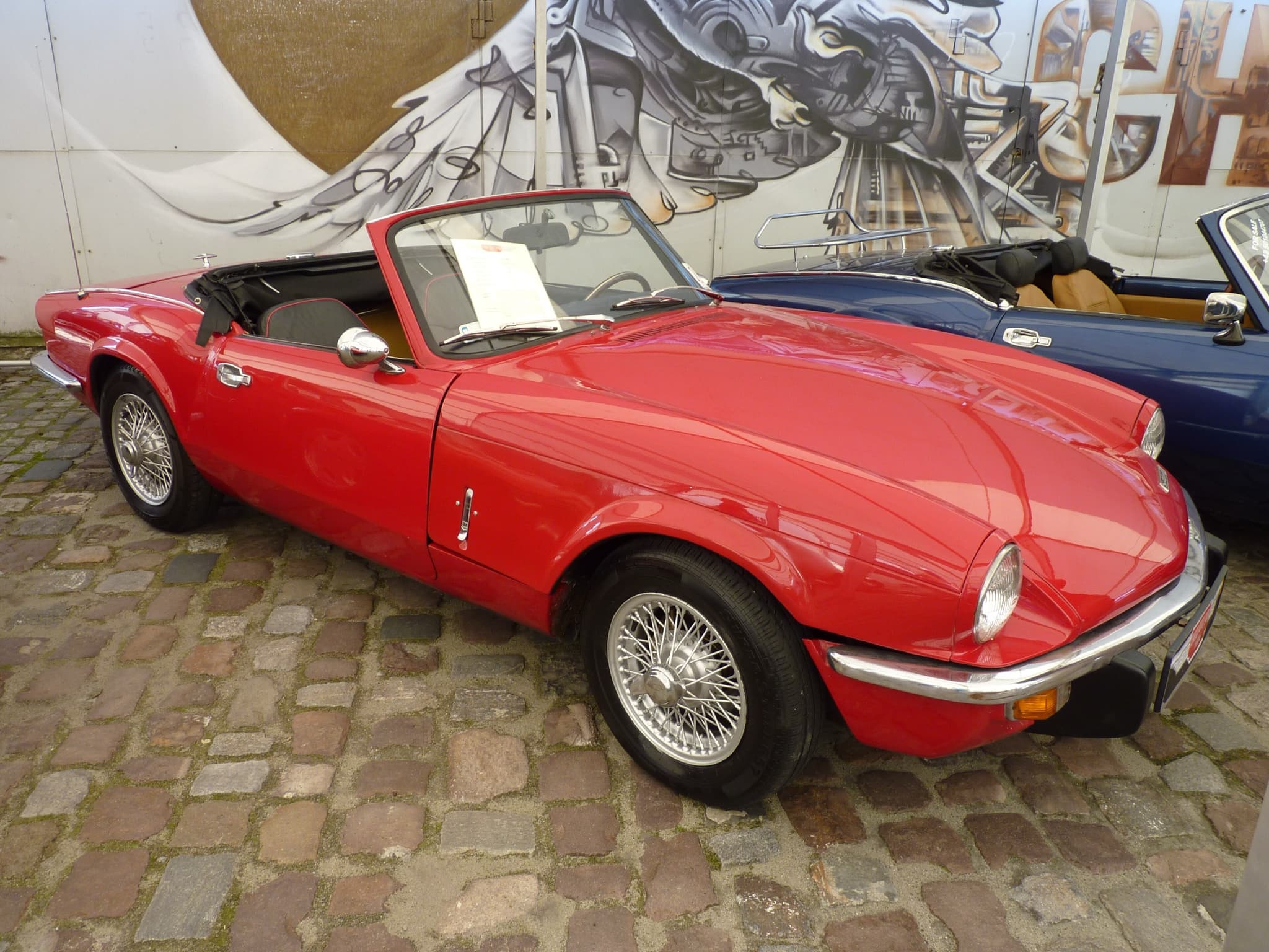 A red Spitfire with its hood folded and wire wheels on cobbles in front of a graffiti mural. Opens a larger view