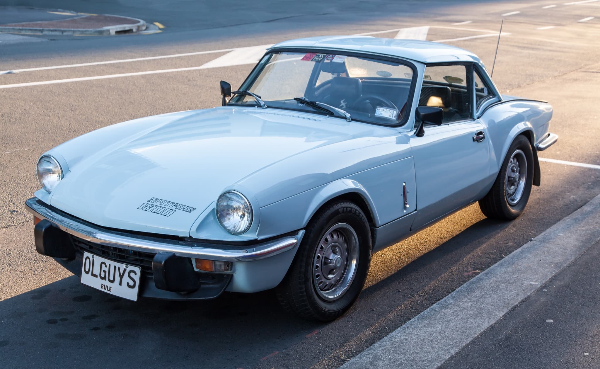 A pale blue Spitfire 1500 with a matching hardtop, parked at a kerb in low evening sun. Opens a larger view