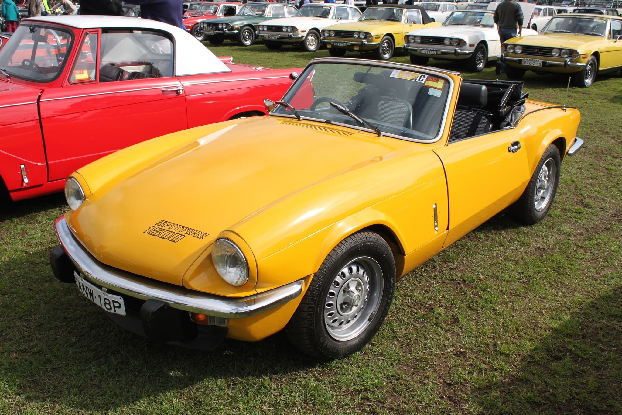 A yellow Spitfire 1500 with its hood folded on a show field, with rows of Triumph Stags behind.