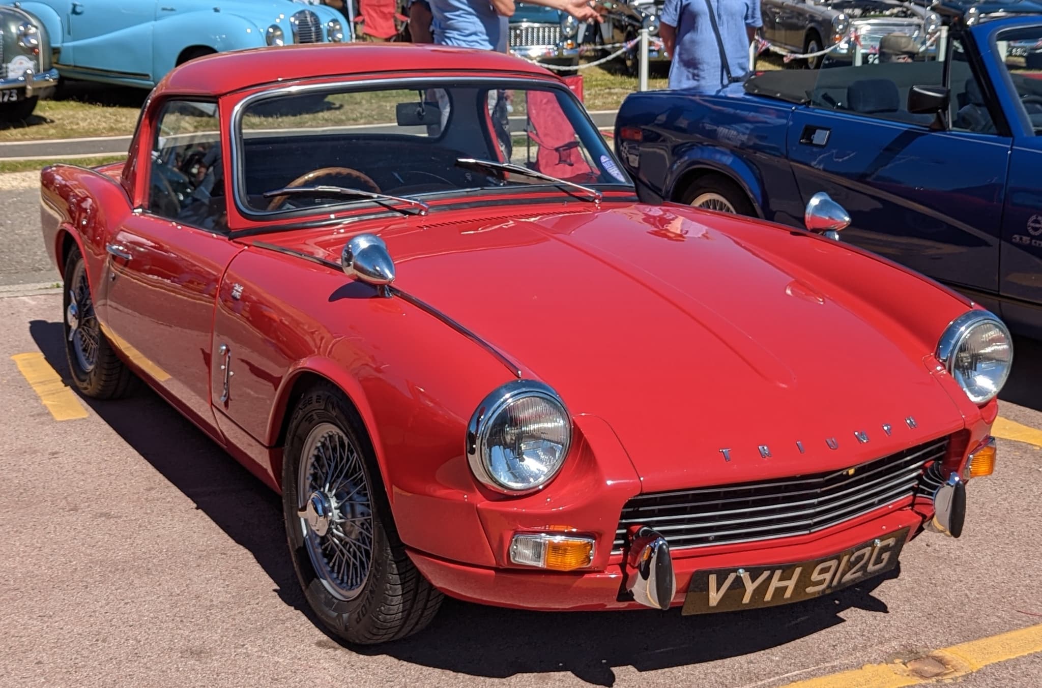 A red Spitfire Mk III with a hardtop and wire wheels, overriders but no front bumper blade, at a show. Opens a larger view