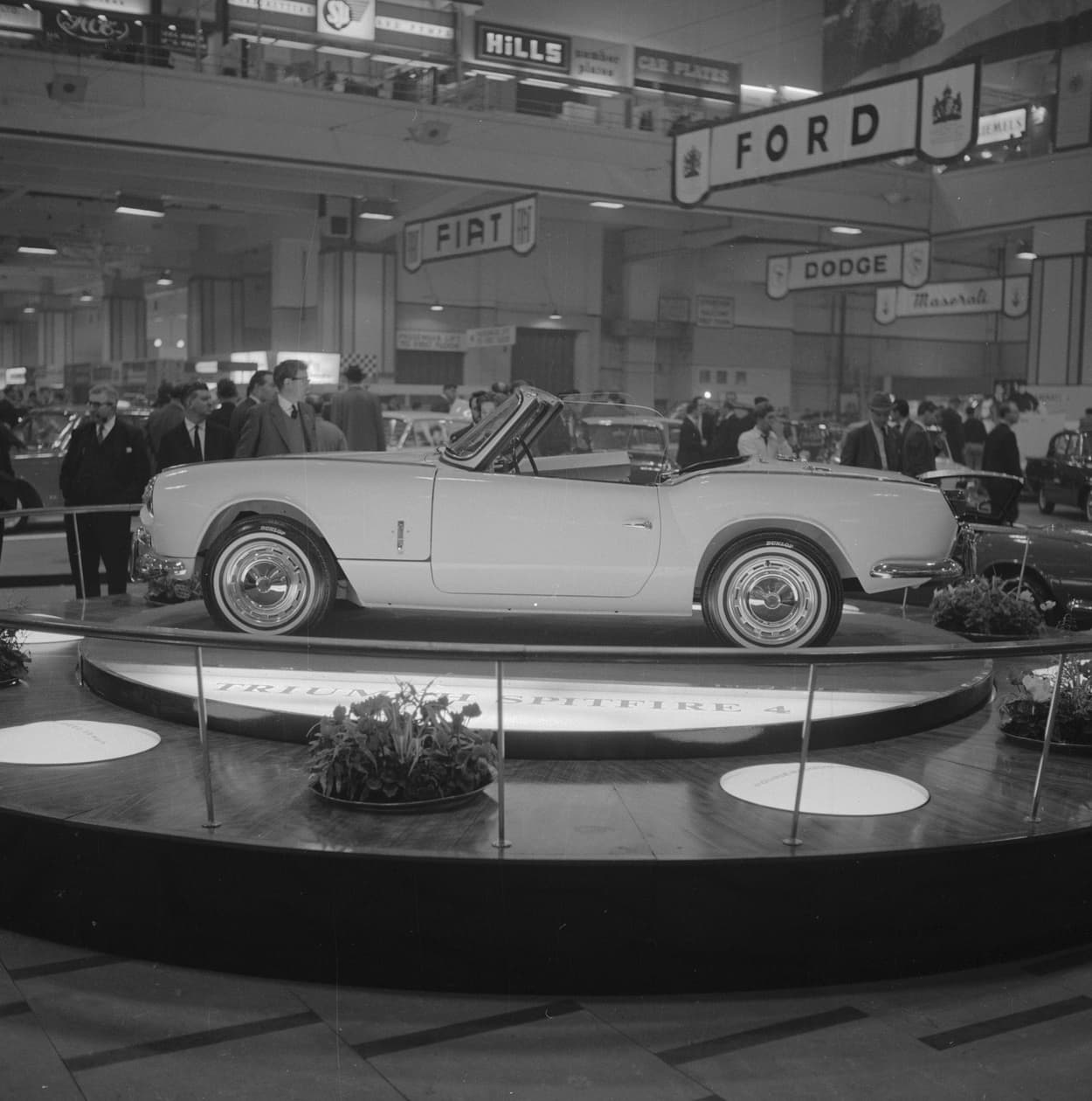 A black-and-white photograph of a Spitfire 4 in profile, hood down, on a lit turntable lettered TRIUMPH SPITFIRE 4, with visitors and stand signs behind.