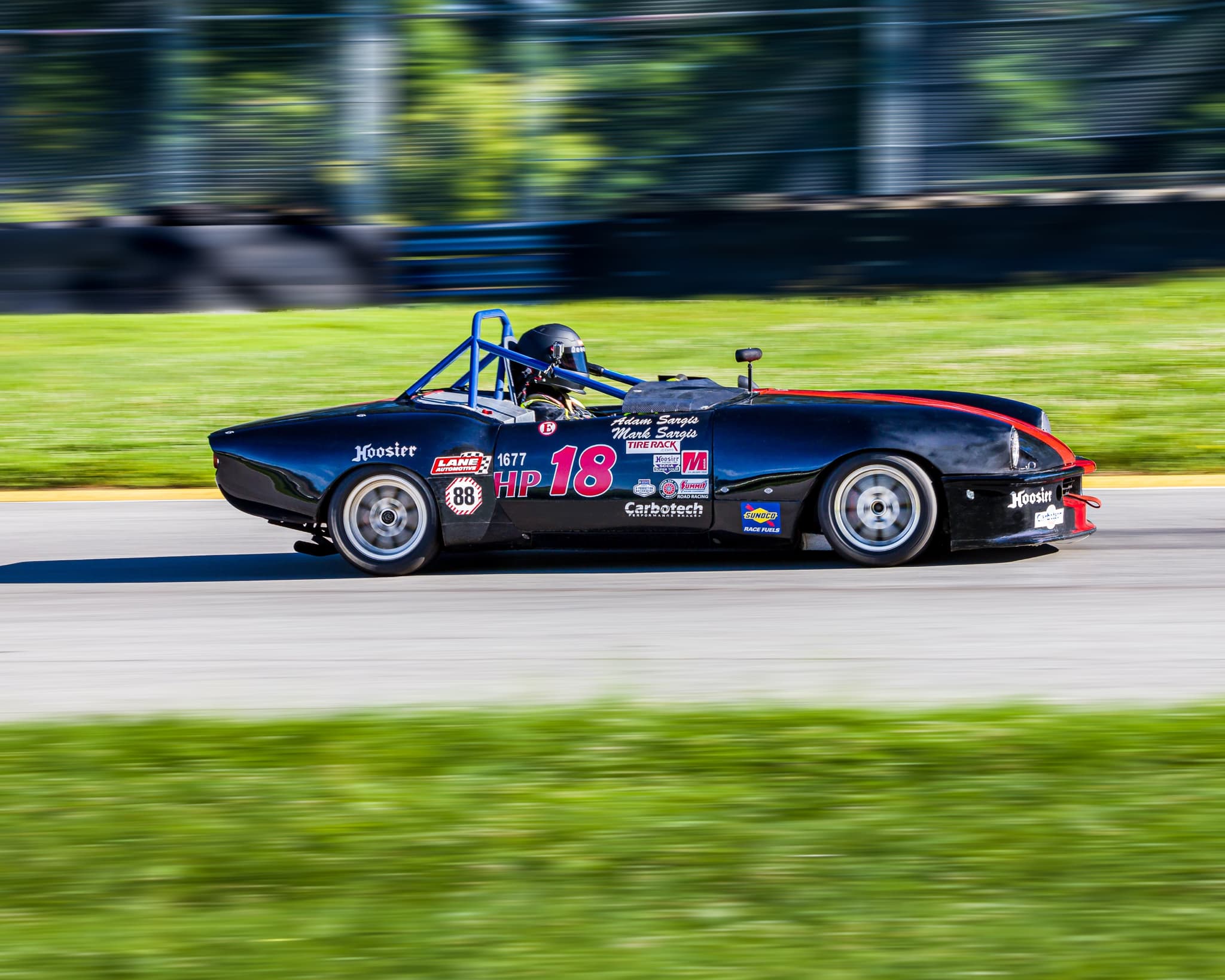 A black Spitfire race car with a blue roll hoop, number 18, panned at speed past a blurred fence. Opens a larger view