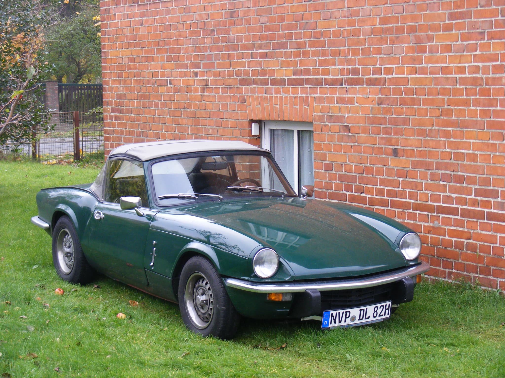 A dark green Spitfire with a beige hood up, parked on grass beside a red-brick house wall. Opens a larger view