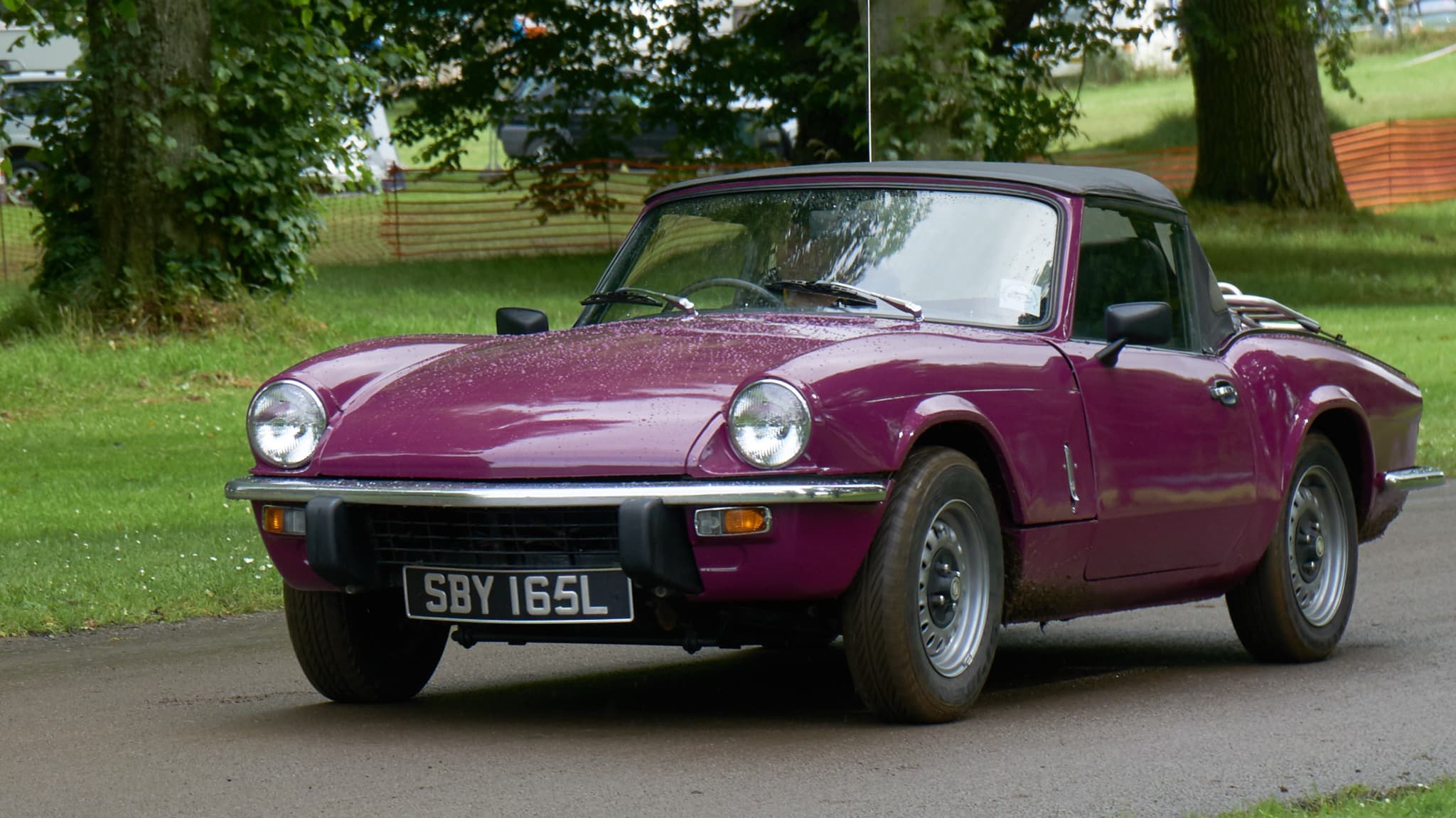 A purple Spitfire with its black hood up on a wet road beside grass and trees. Opens a larger view