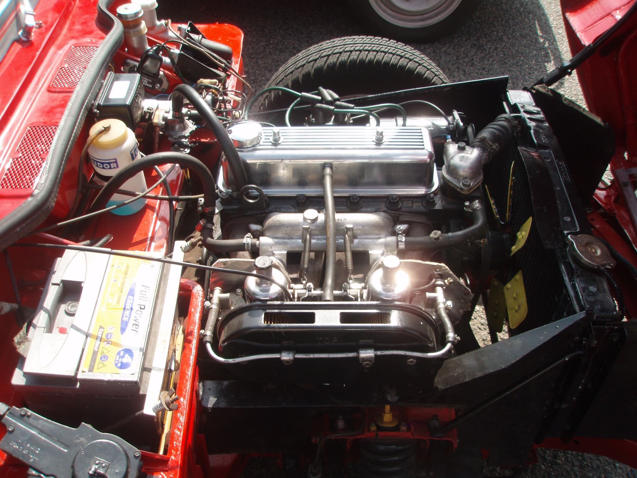 An open engine bay with twin carburettors, a polished rocker cover, a battery and the radiator.
