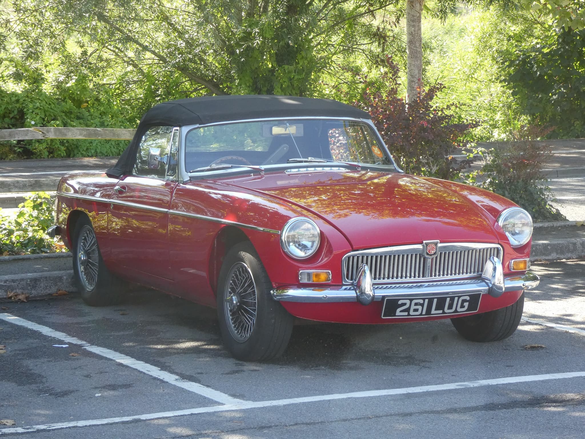 A red sports car with a black hood up, chrome bumpers, a vertical-slat grille and wire wheels, parked in a car park under trees. Opens a larger view