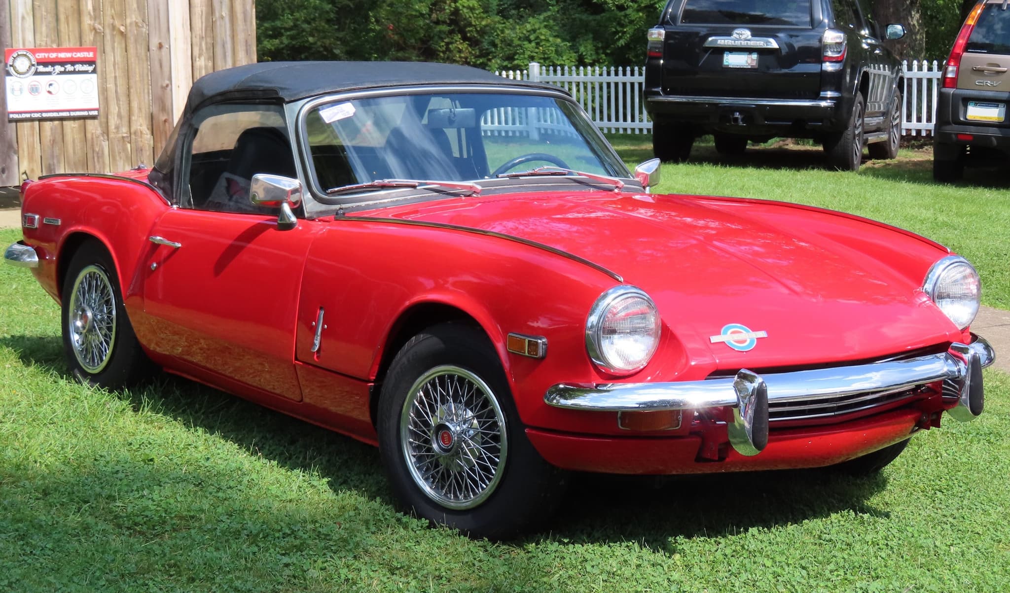 A red US-market Spitfire with its black hood up, wire wheels and a roundel badge, parked on grass. Opens a larger view